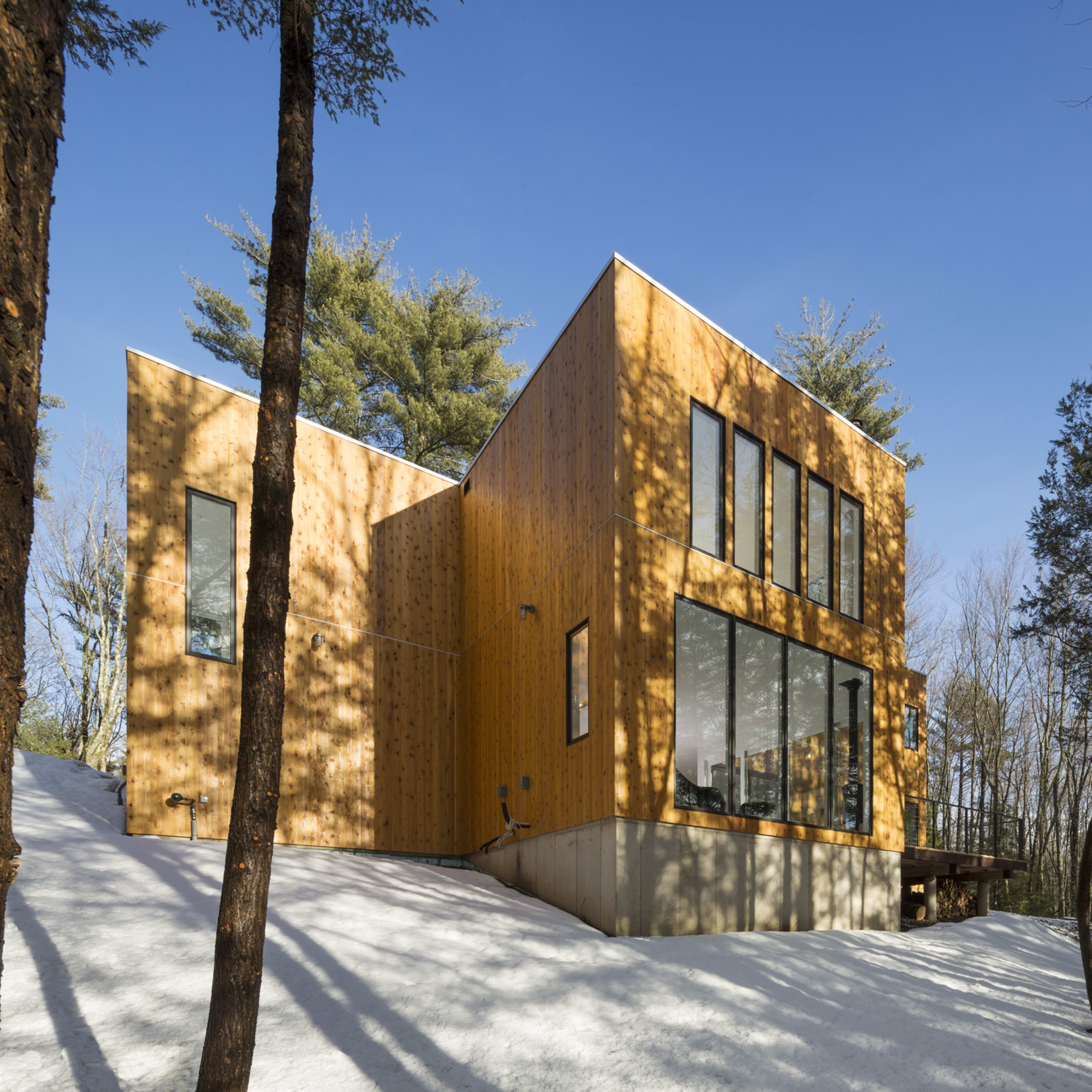 A modern wooden house with large windows is surrounded by trees and snow-covered ground.