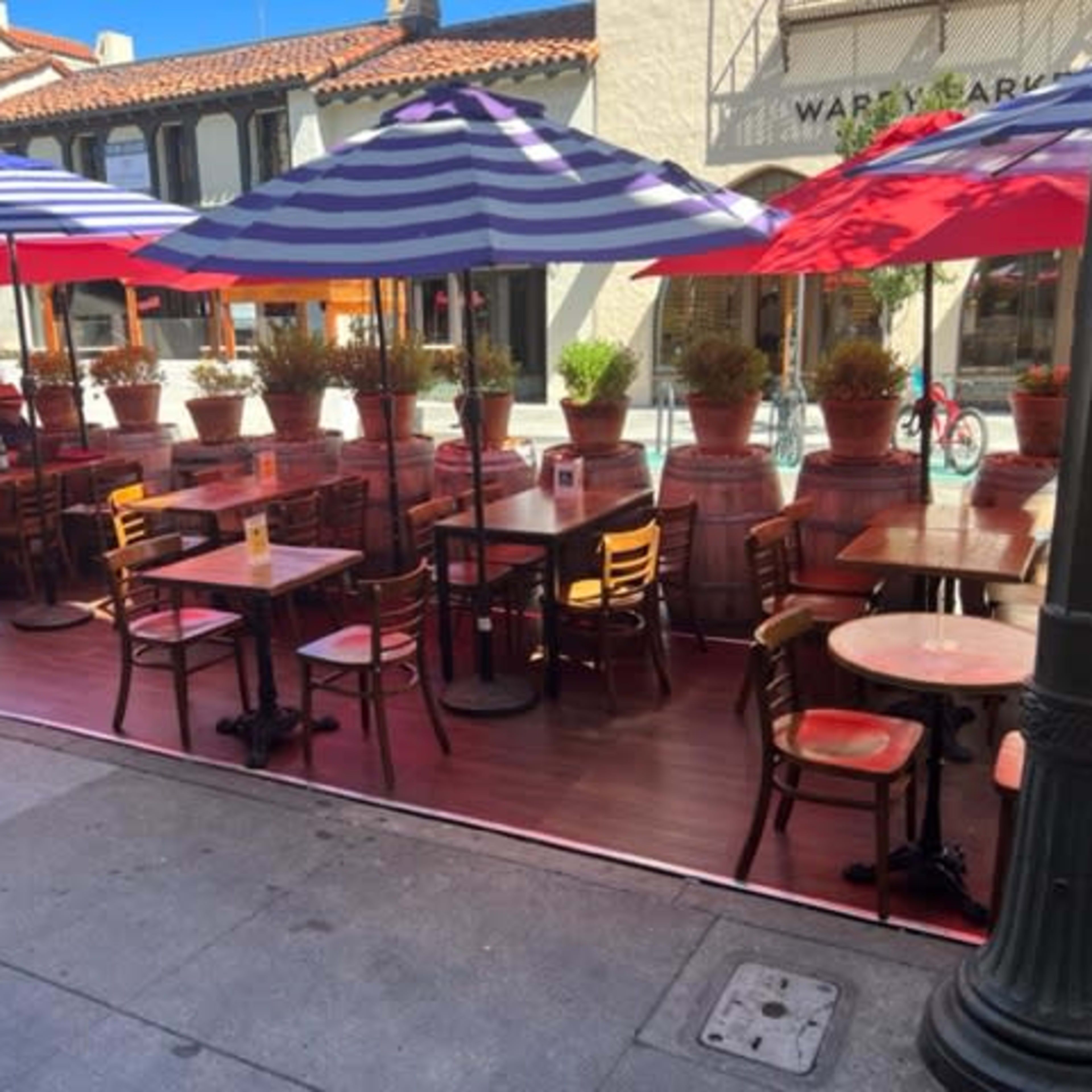 A row of outdoor tables and chairs, shaded by striped umbrellas, is set up outside a restaurant with potted plants along the perimeter.
