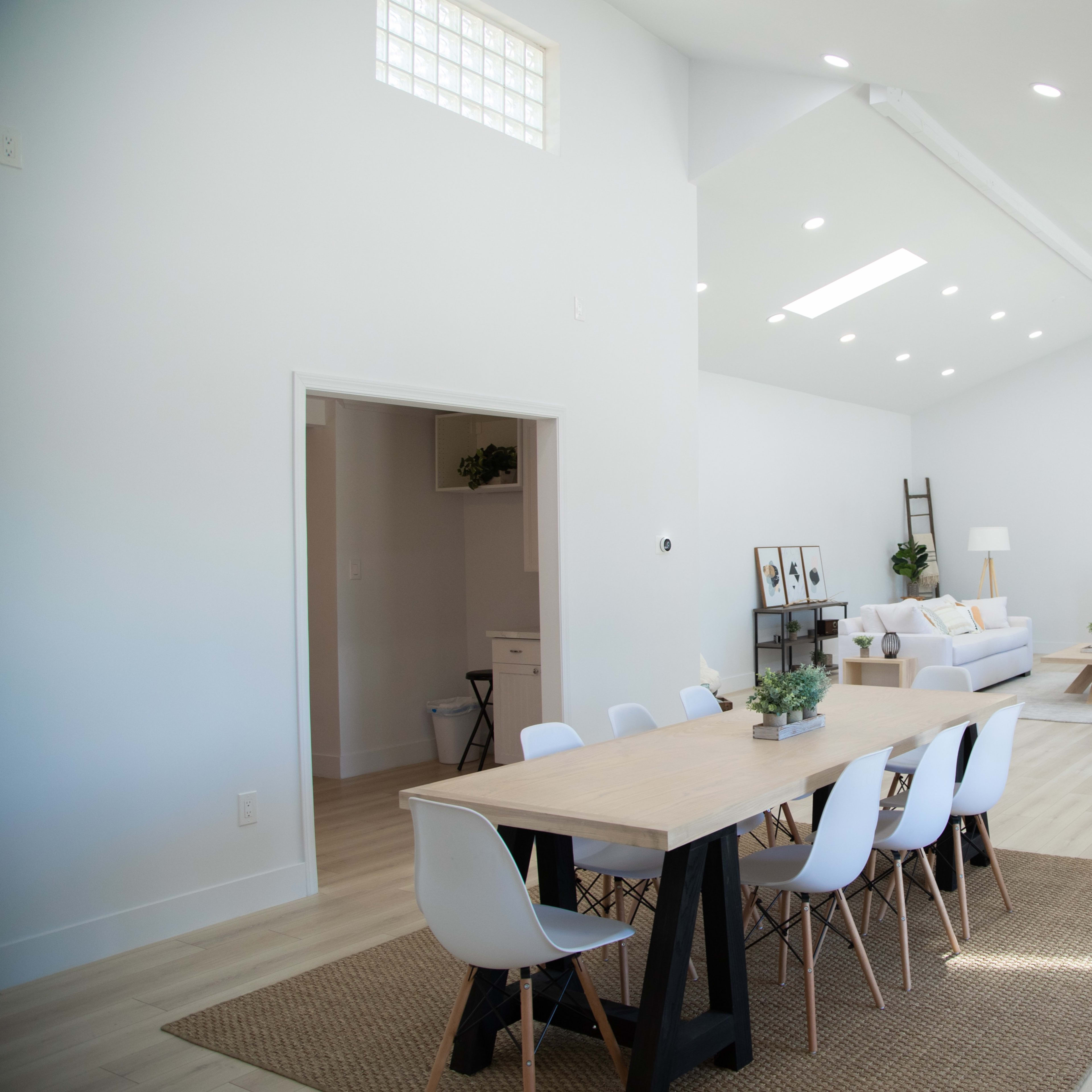 A spacious, brightly lit interior of a modern living area and dining space with a wooden table surrounded by white chairs, a seating area with a sofa, and large windows.