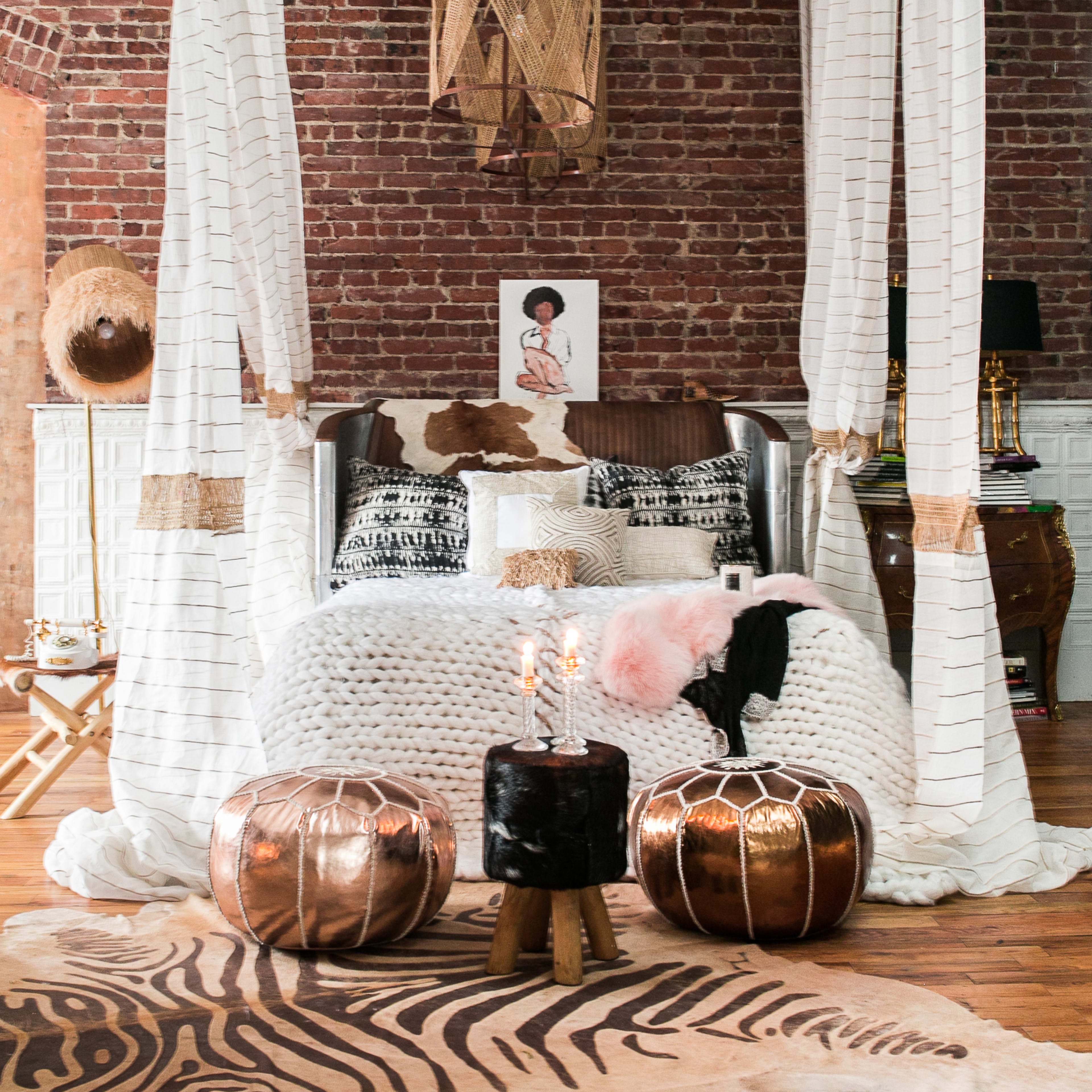 A cozy bedroom features a large bed draped with white fabric, surrounded by decorative pillows, a patterned rug, and two metallic poufs.
