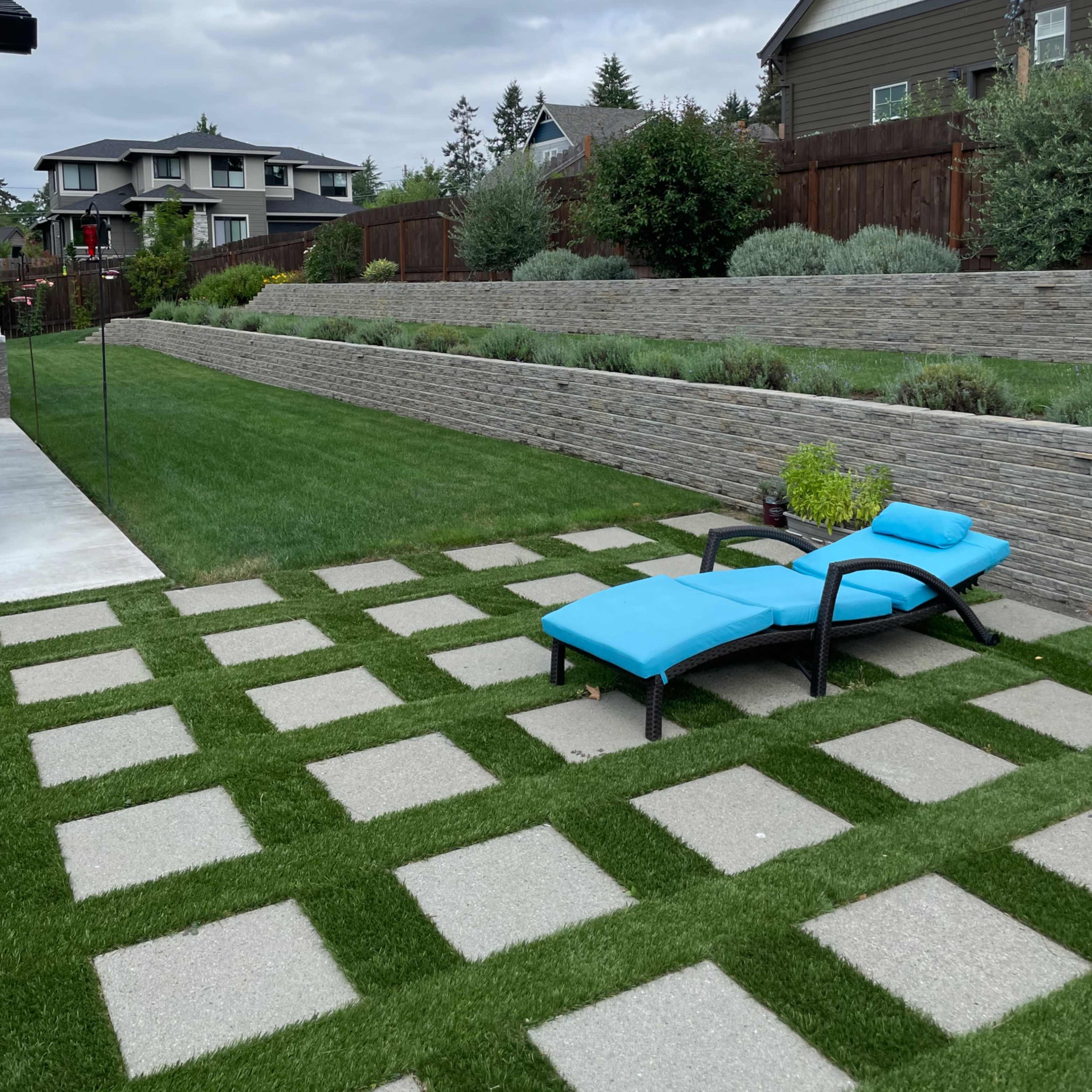 A landscaped backyard with a concrete paver pathway, a blue lounge chair, and neatly trimmed grass bordered by stone walls and shrubs.
