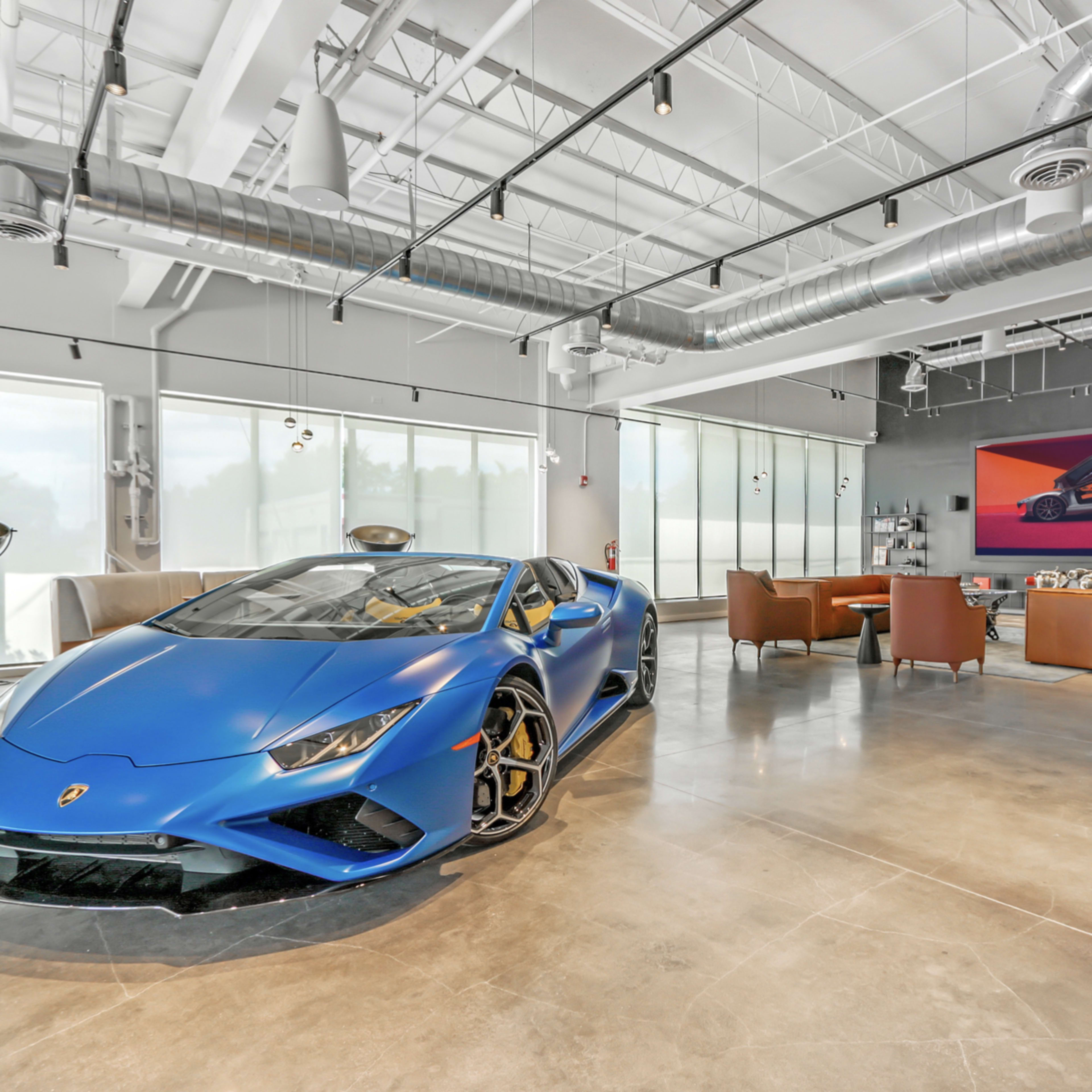 A blue Lamborghini is prominently displayed in a modern showroom with large windows and contemporary seating.