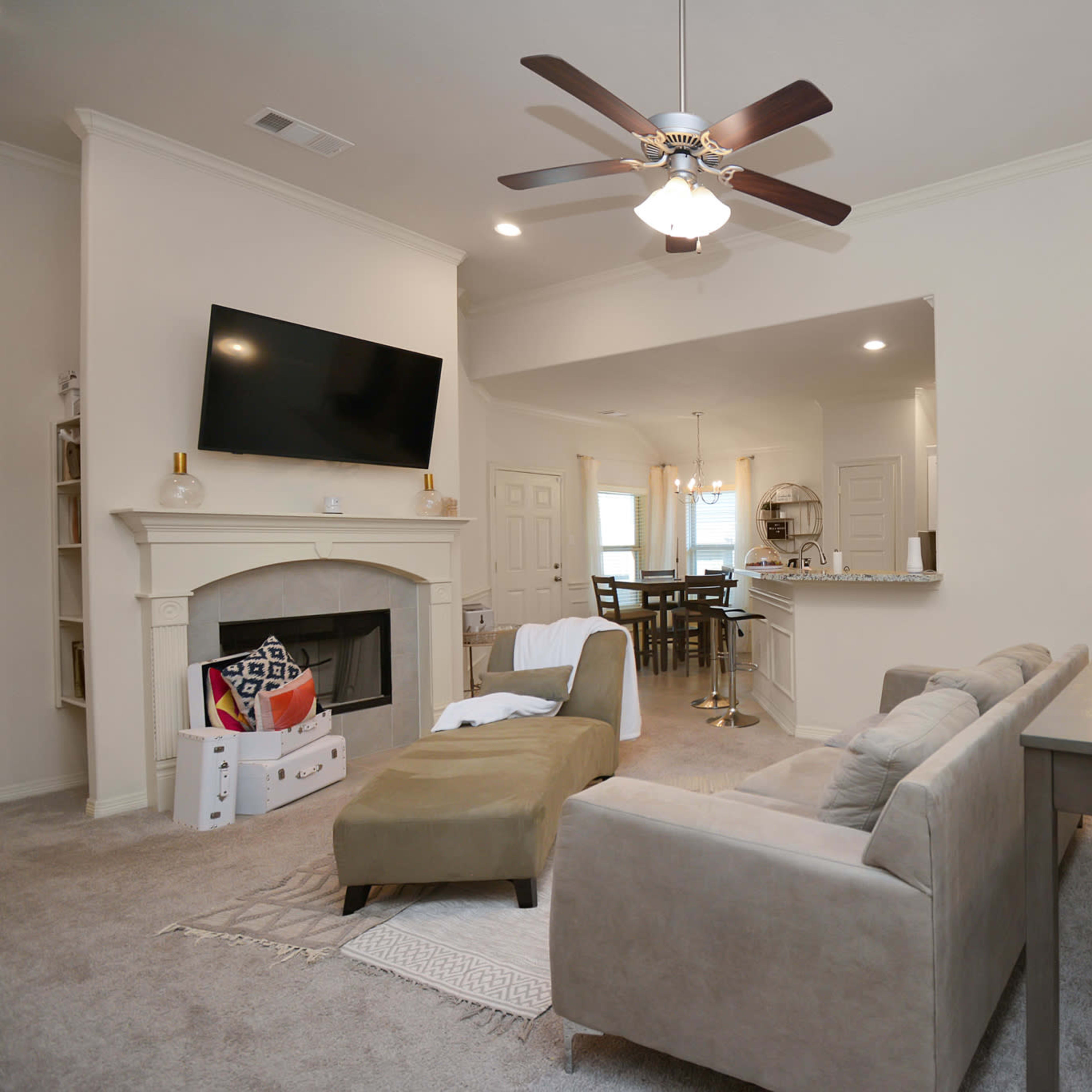 A living room with a beige sofa, a mounted television above a fireplace, and a dining area visible in the background.