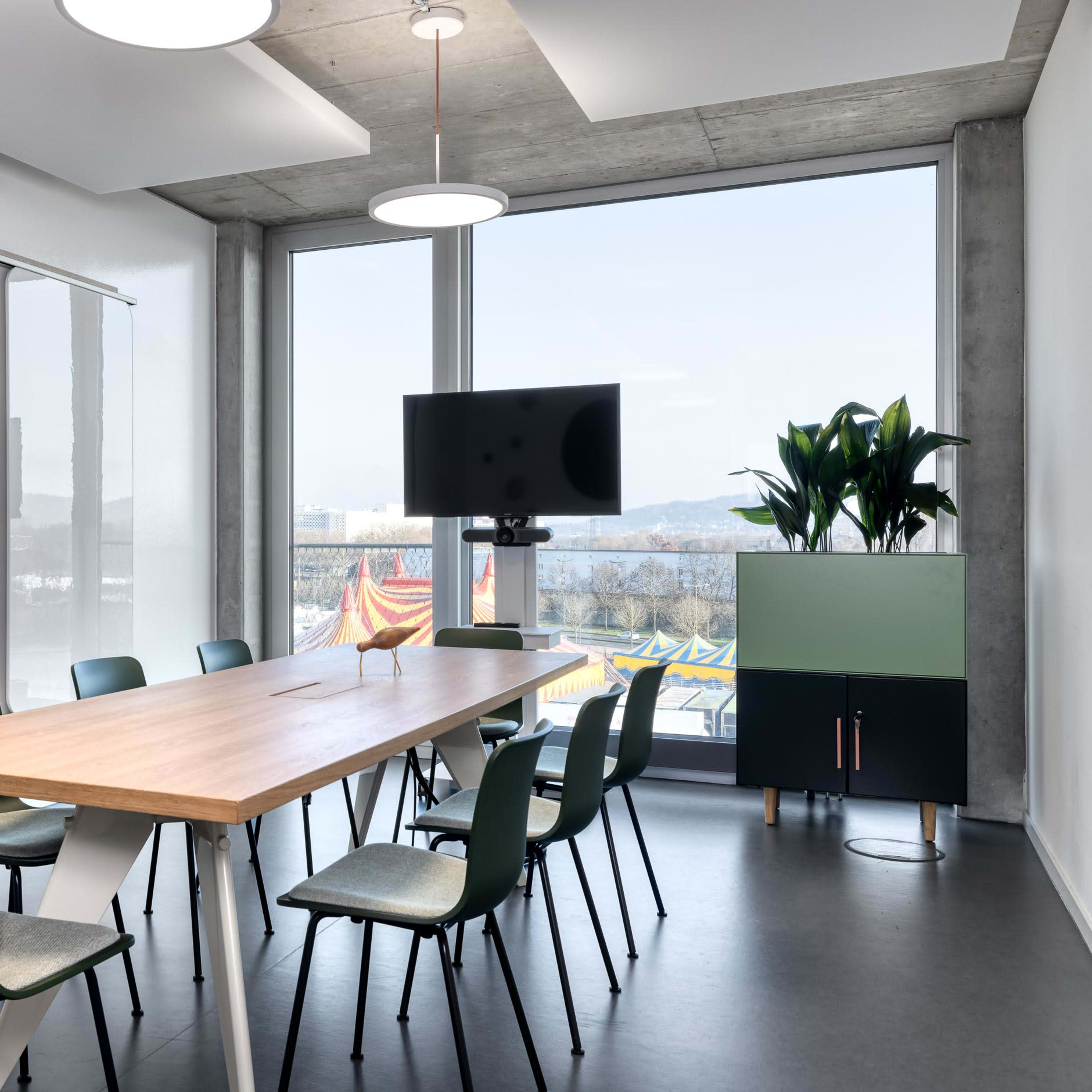 A modern conference room with a long wooden table surrounded by green chairs, a television on the wall, and large windows overlooking an outdoor area.