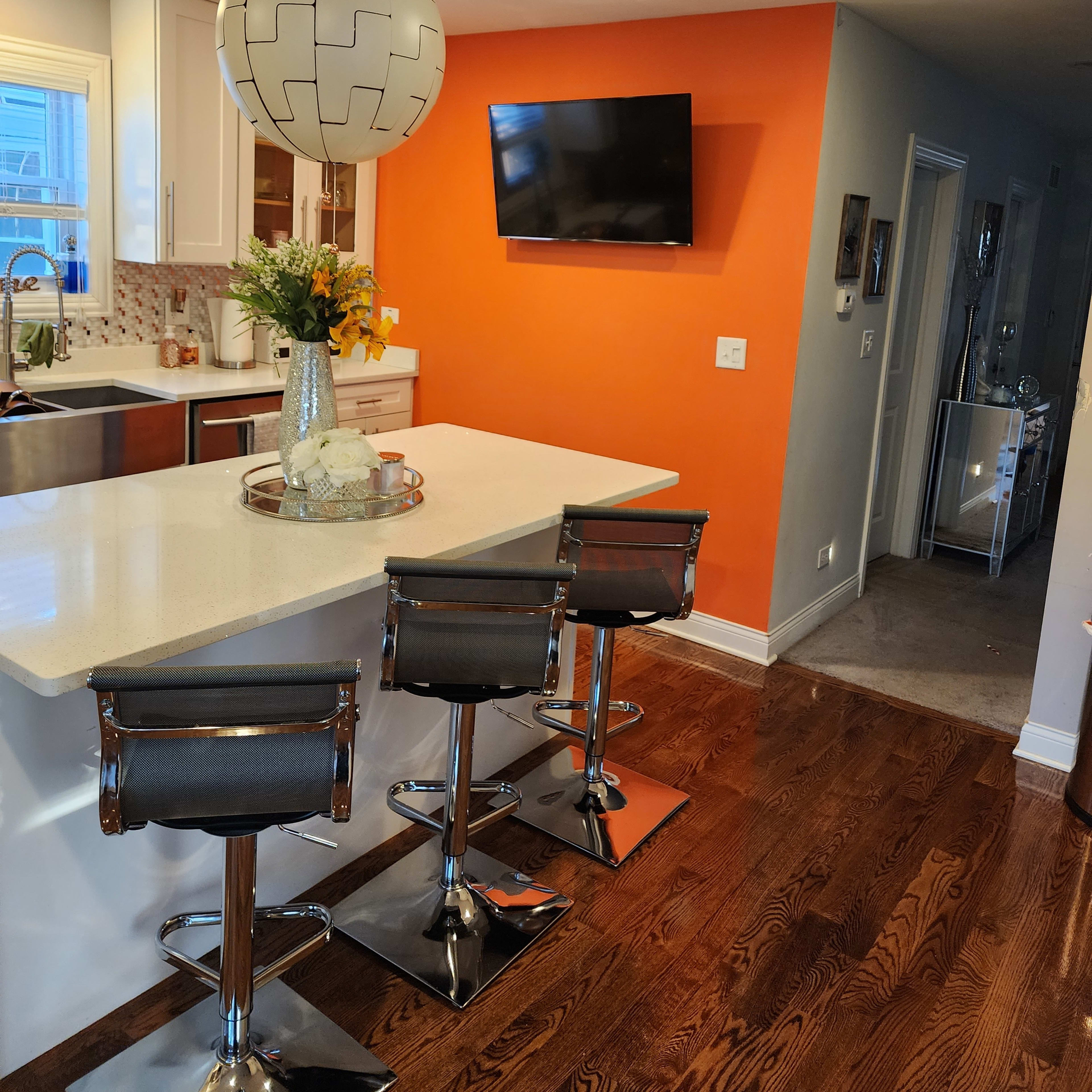 A modern kitchen featuring a white countertop with three bar stools, an orange accent wall, and a television mounted above the countertop.