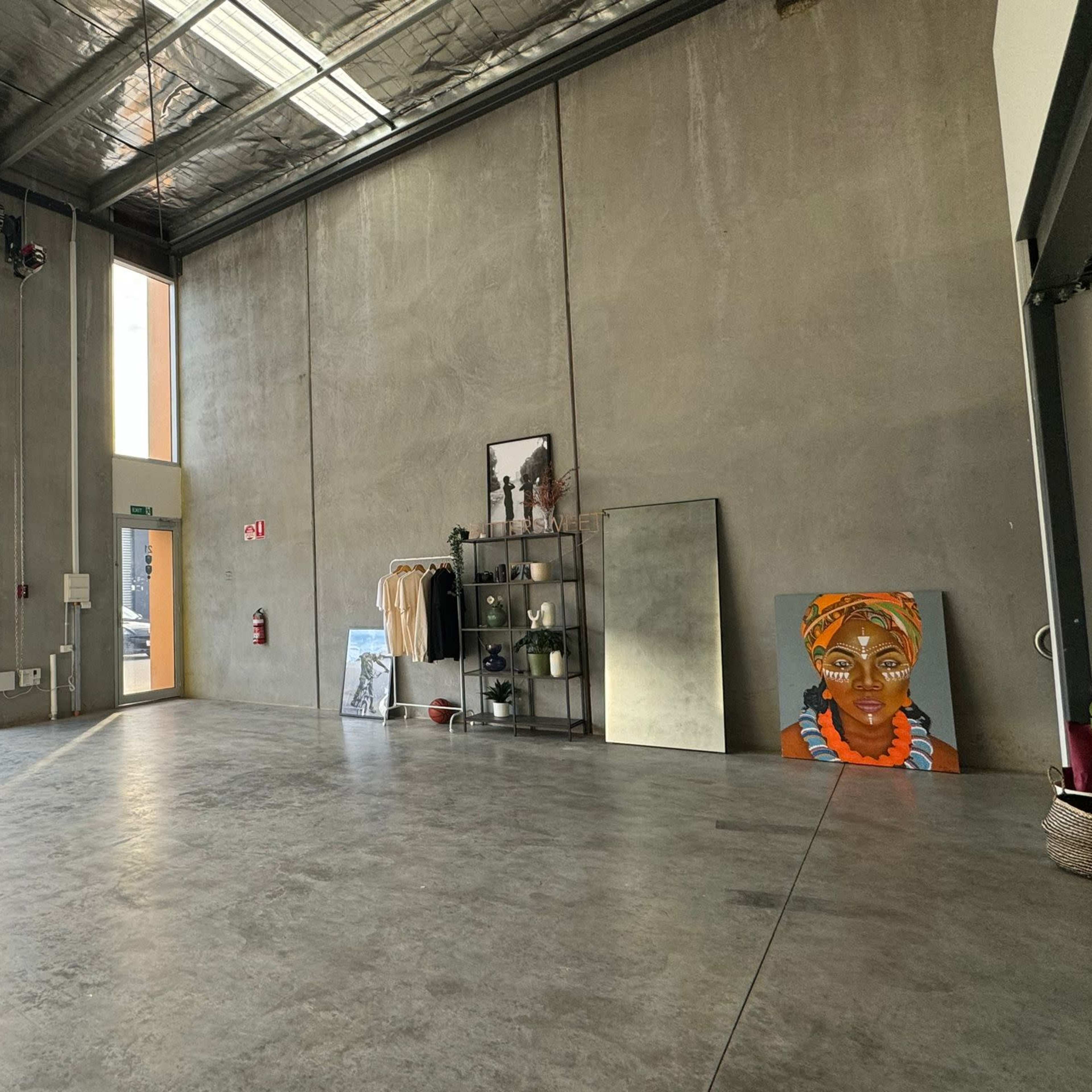 A spacious, bare concrete interior of a building with a large roll-up door, a display shelf holding clothes and decor, a standing mirror, and a colorful artwork of a woman on the floor.