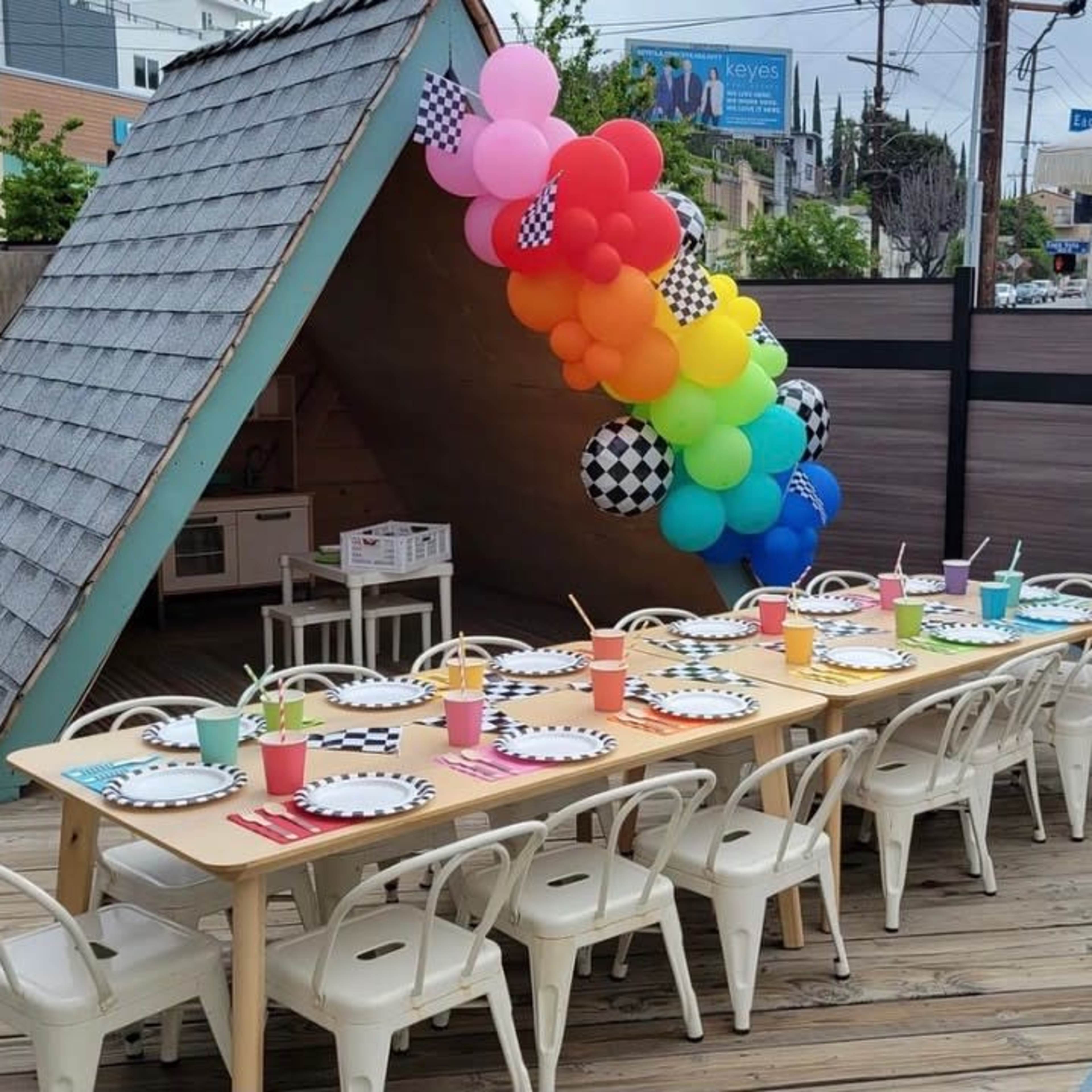 A long table is set with colorful cups and checkered plates, positioned outside near a small playhouse adorned with rainbow balloons and a checkered banner.