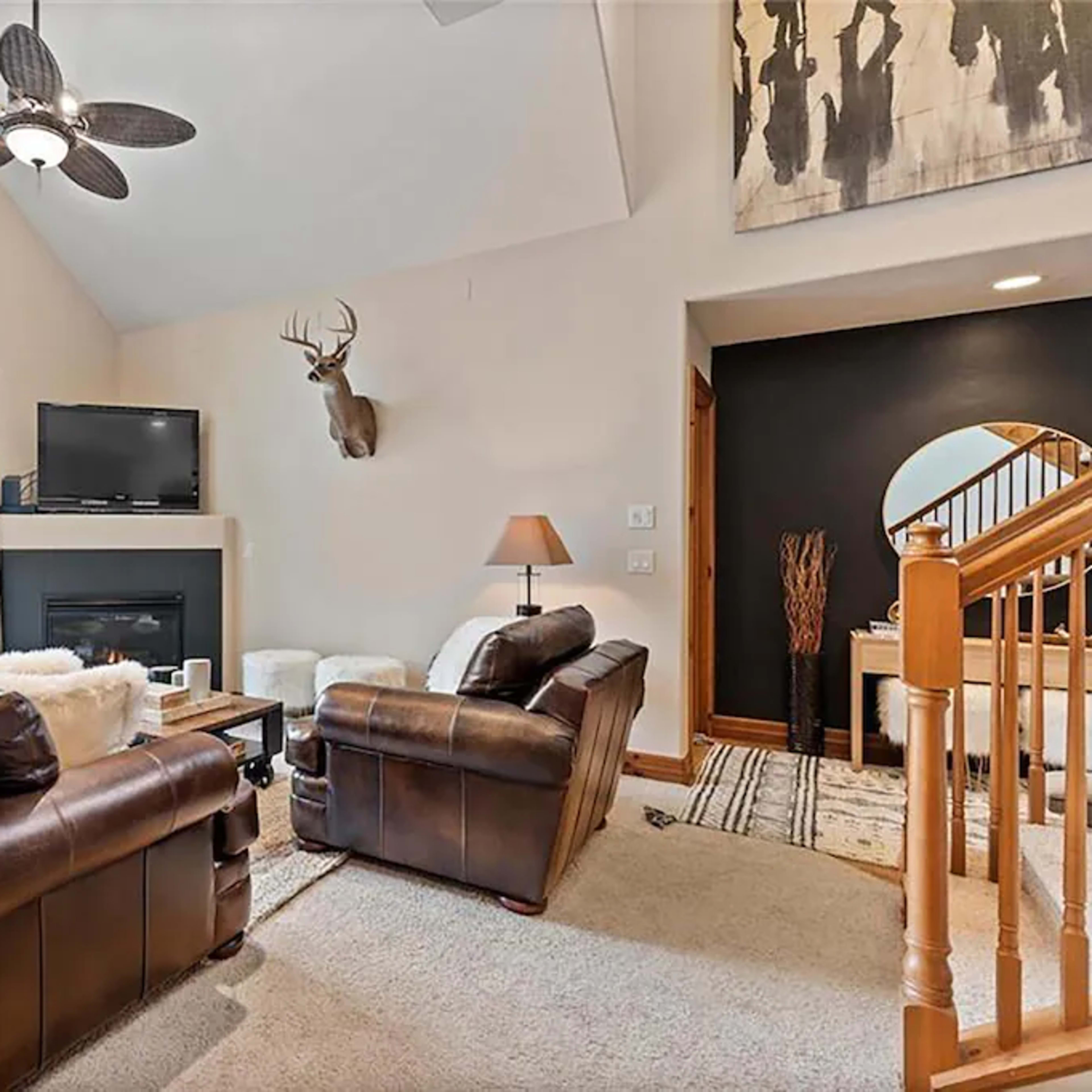 The living room features leather sofas, a mounted deer head on the wall, a television above a modern fireplace, and large windows letting in natural light.