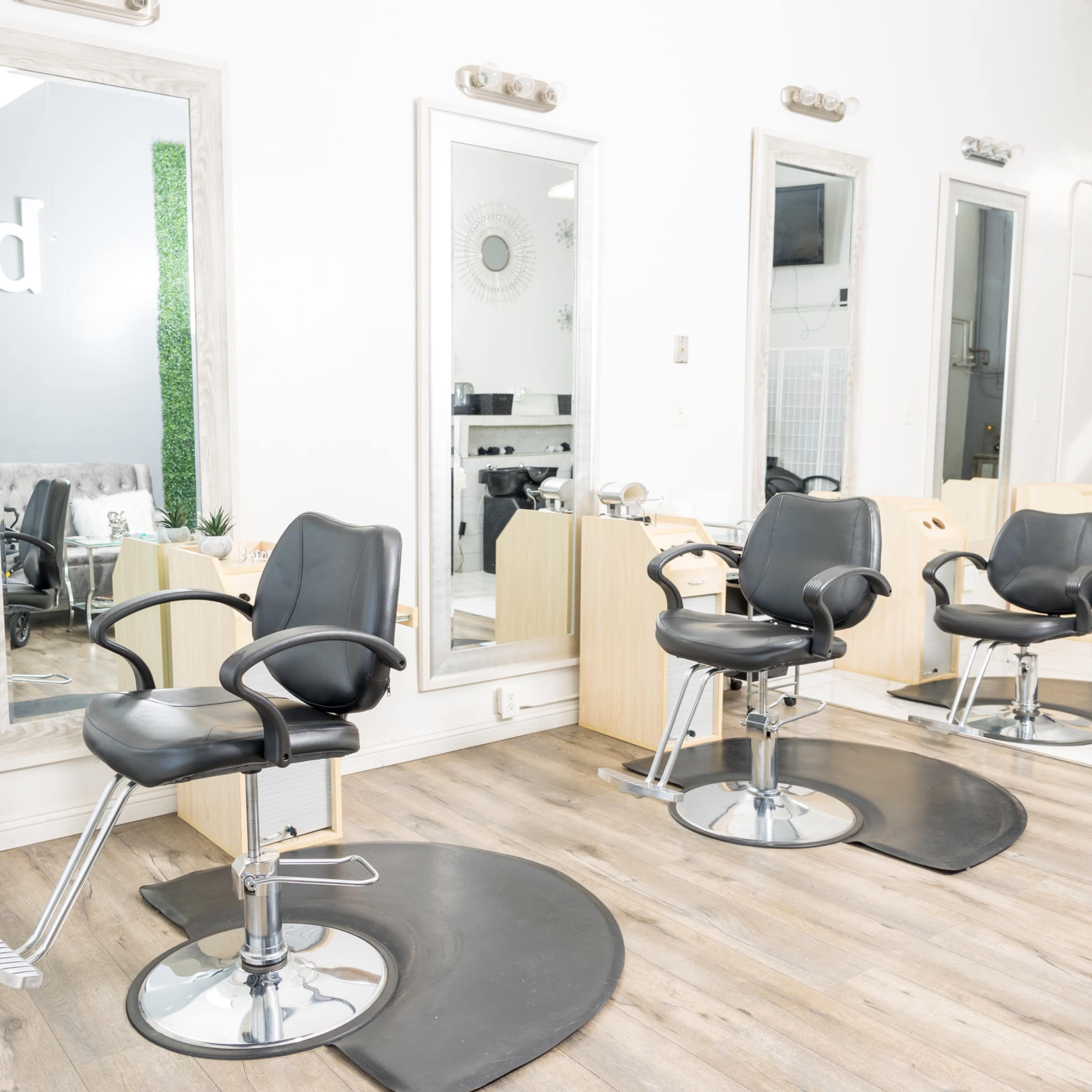 A hair salon interior with two black styling chairs, large mirrors, and wooden flooring.