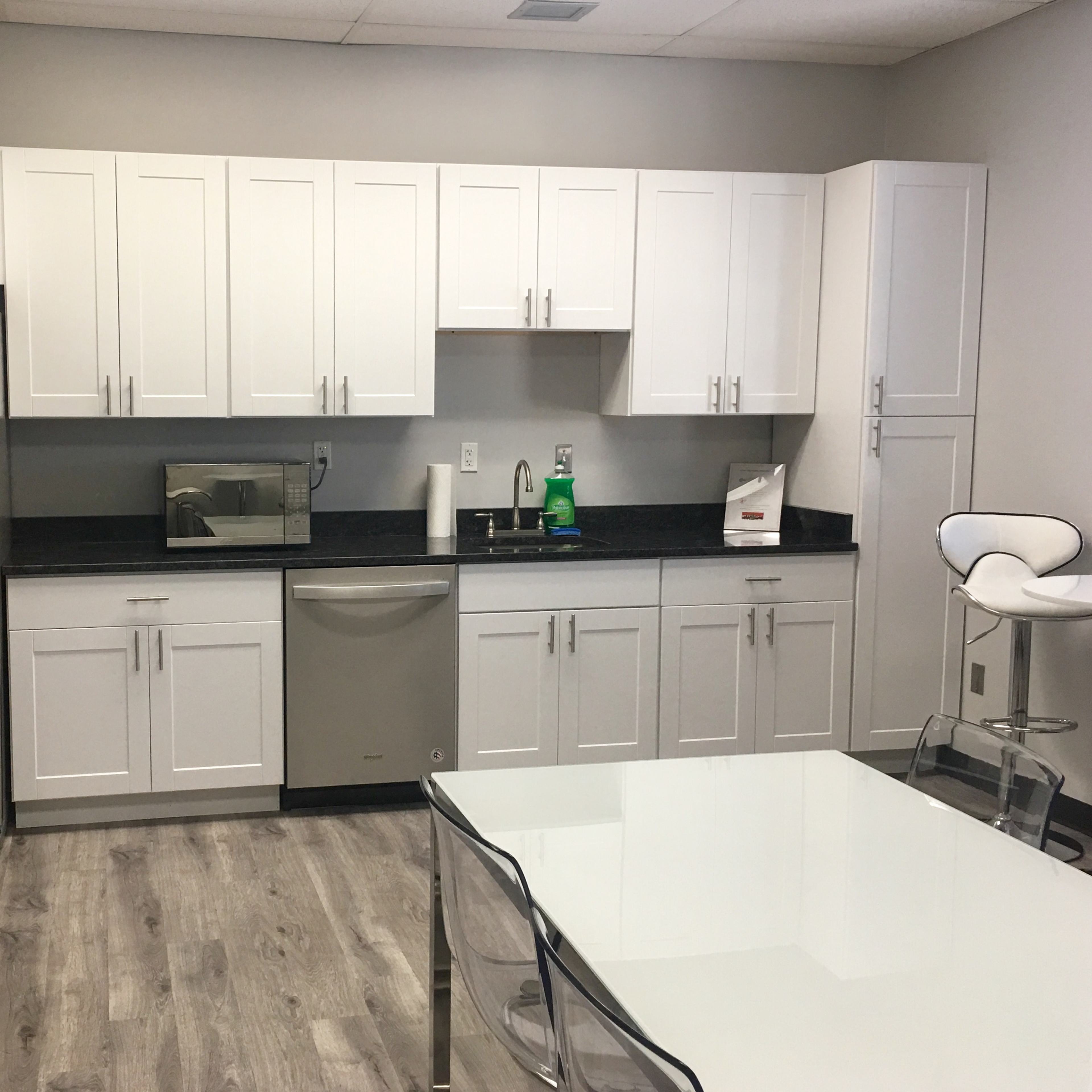 A modern kitchen with white cabinetry, a stainless steel refrigerator, a dishwasher, and a clear table with chairs.
