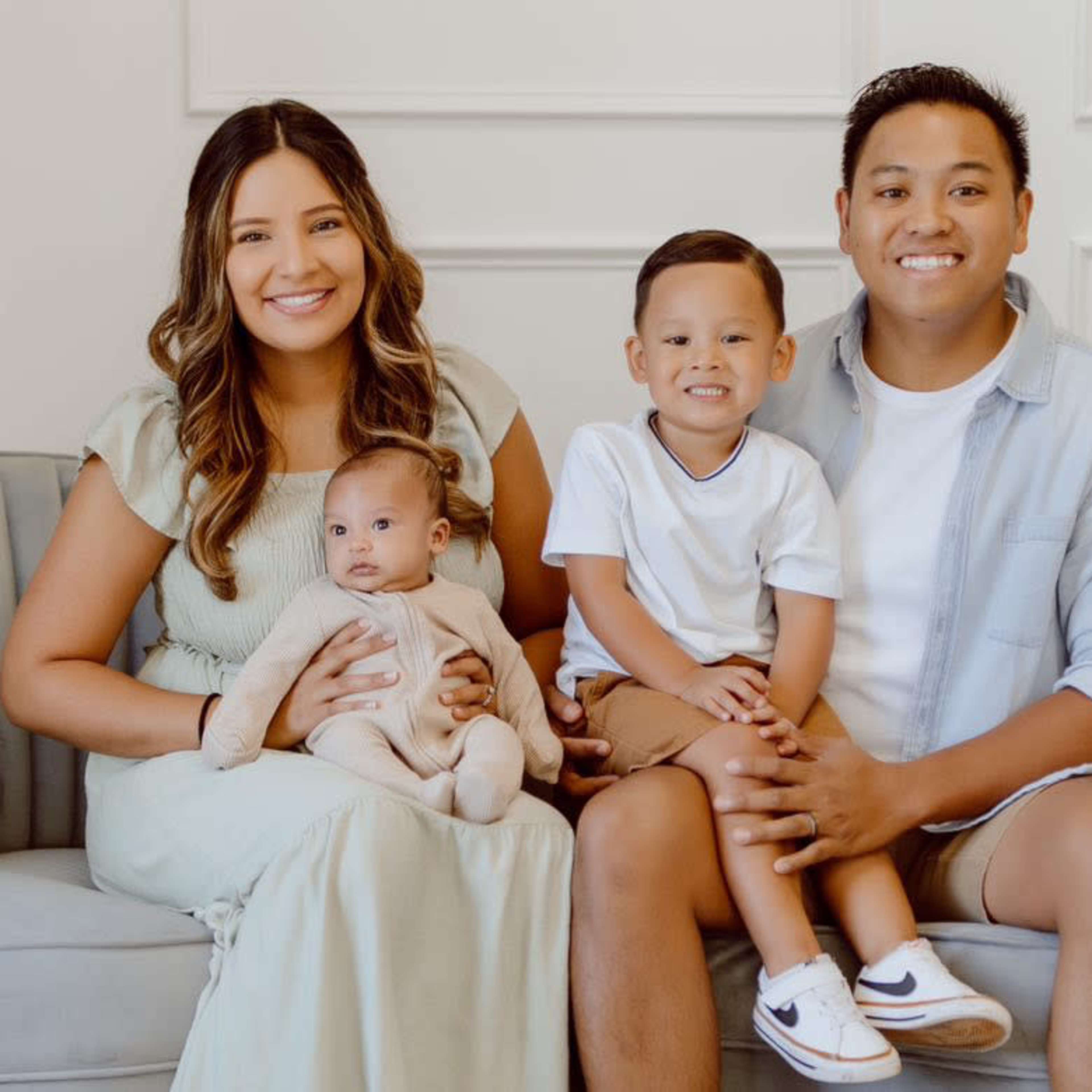 A family of four is seated on a gray couch, with the mother holding a baby and the father beside her, while their young son sits next to them.