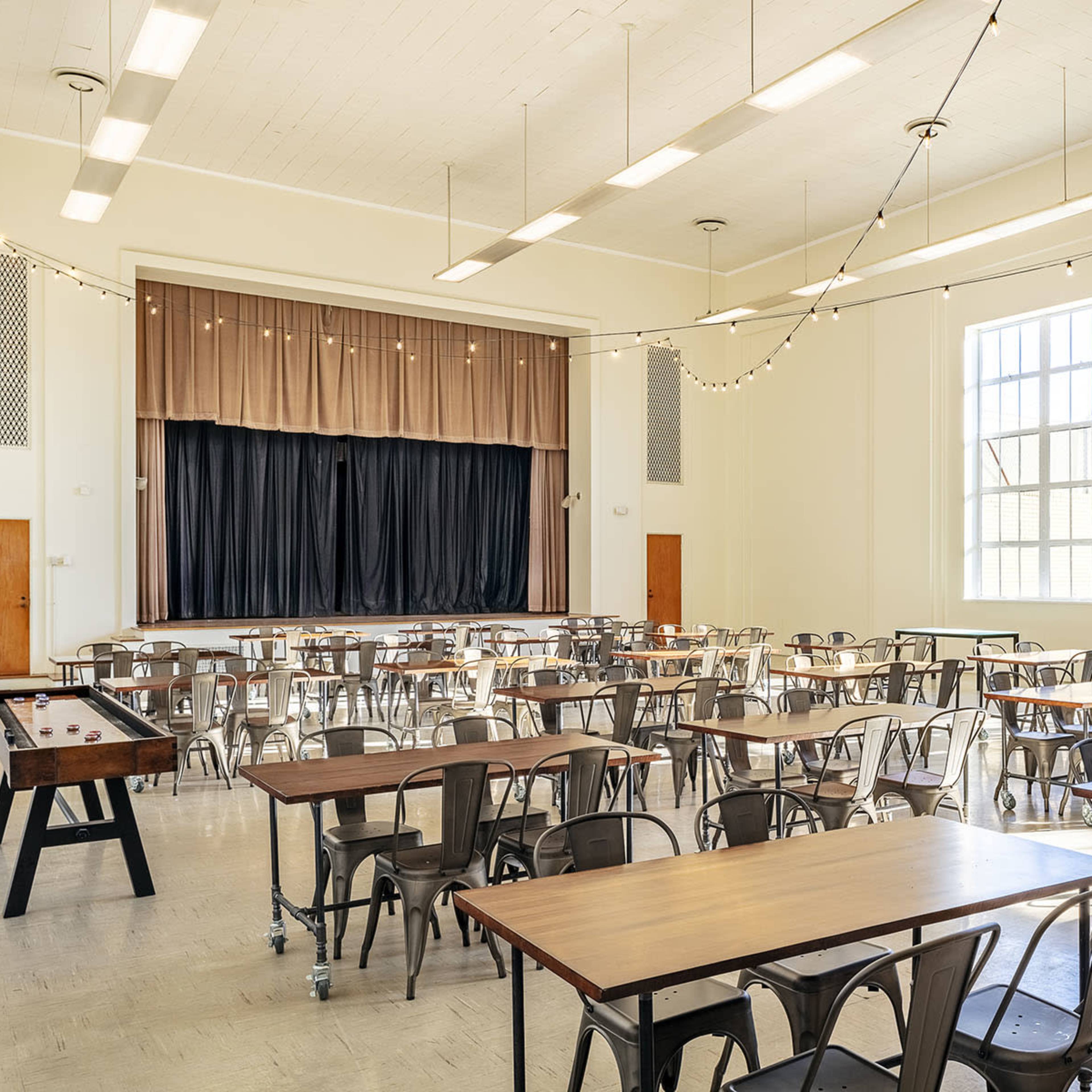 A spacious indoor hall with rows of tables and chairs arranged facing a stage at the front, illuminated by large windows.
