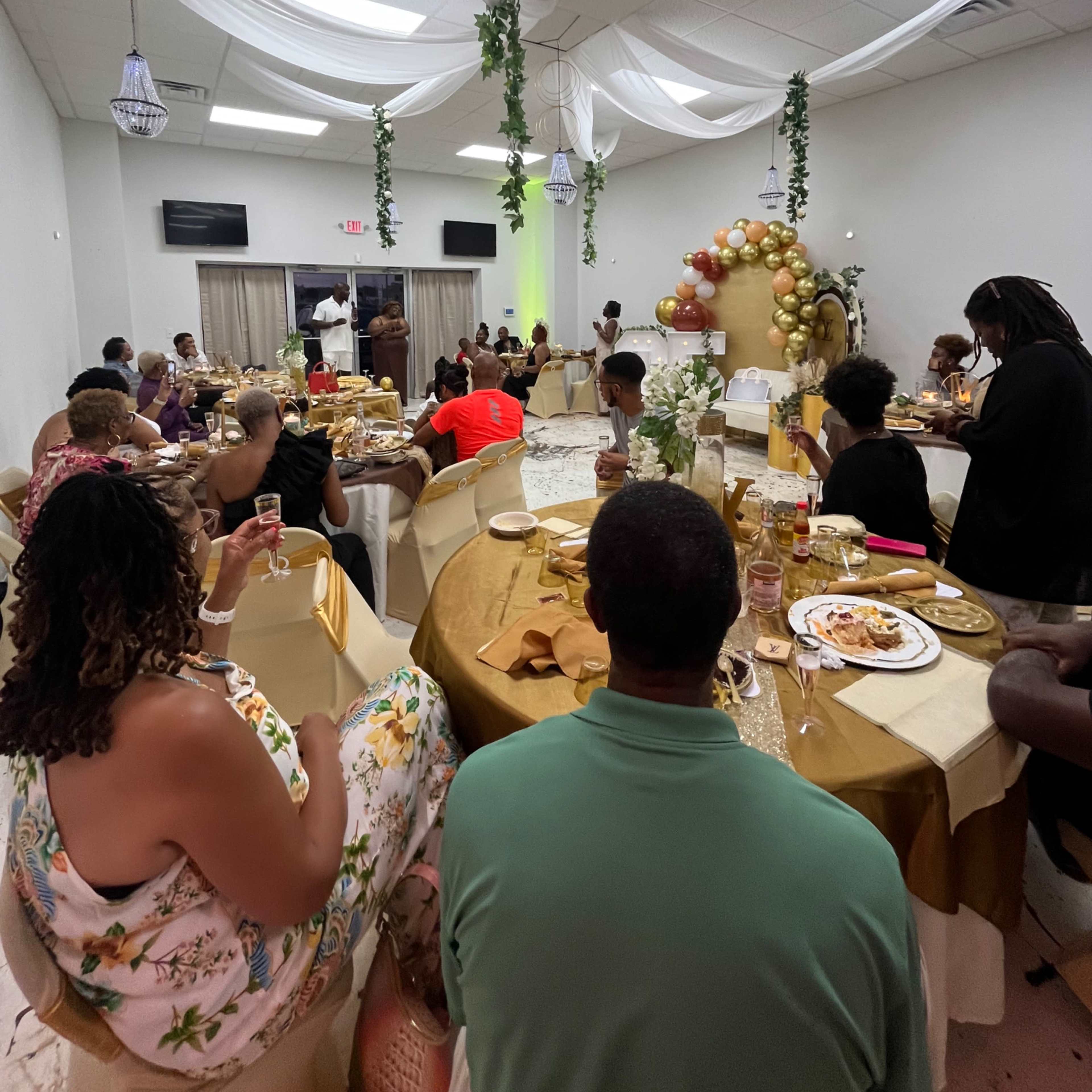 A gathering of guests seated at tables in a decorated hall, enjoying food and drinks during a celebratory event.