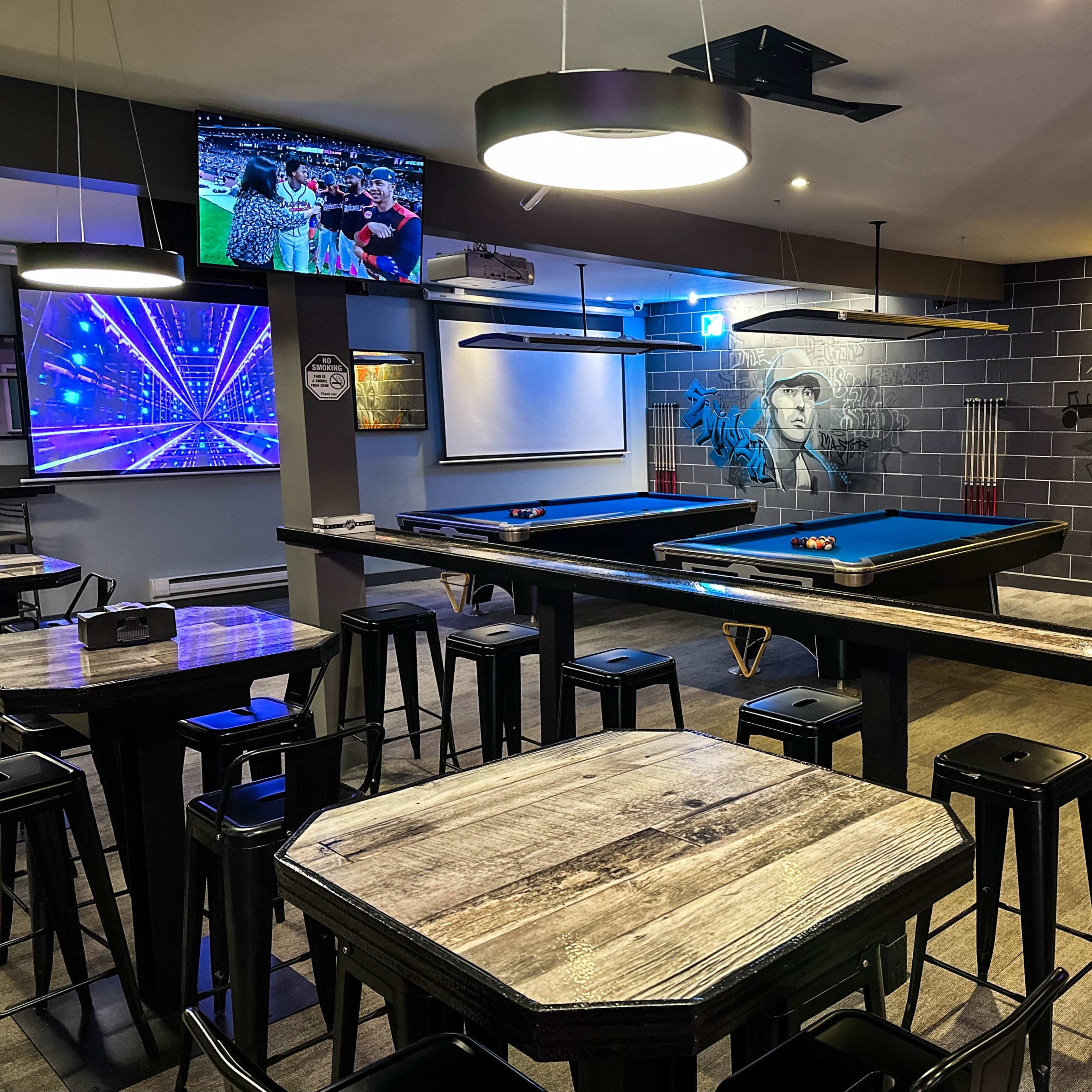 A billiard room featuring several pool tables, bar stools, and a large screen displaying visuals, with wall murals of figures and vibrant artwork.