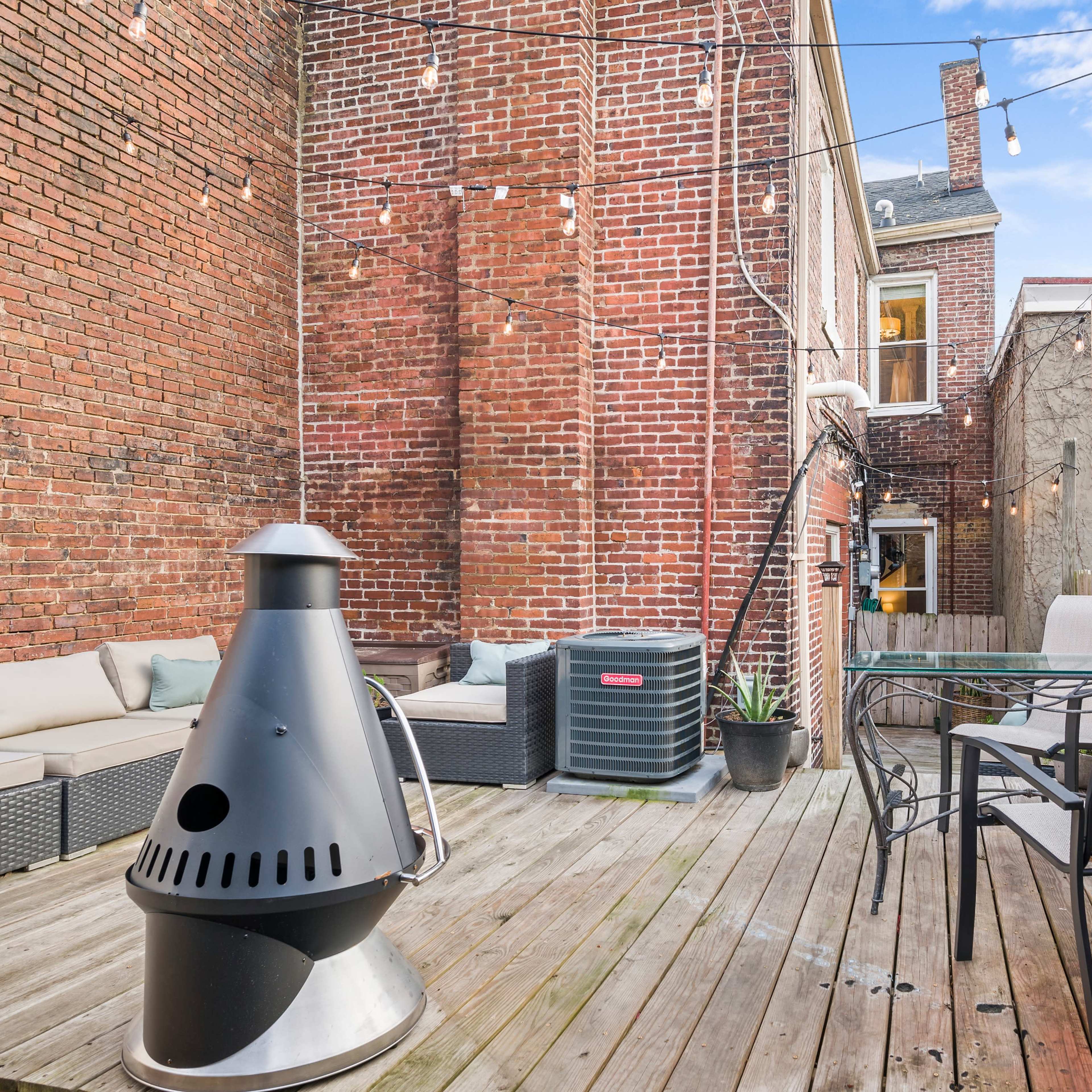 A wooden deck in an outdoor space with a seating area, a metal chiminea, and a small dining table, surrounded by brick walls and a wooden fence.