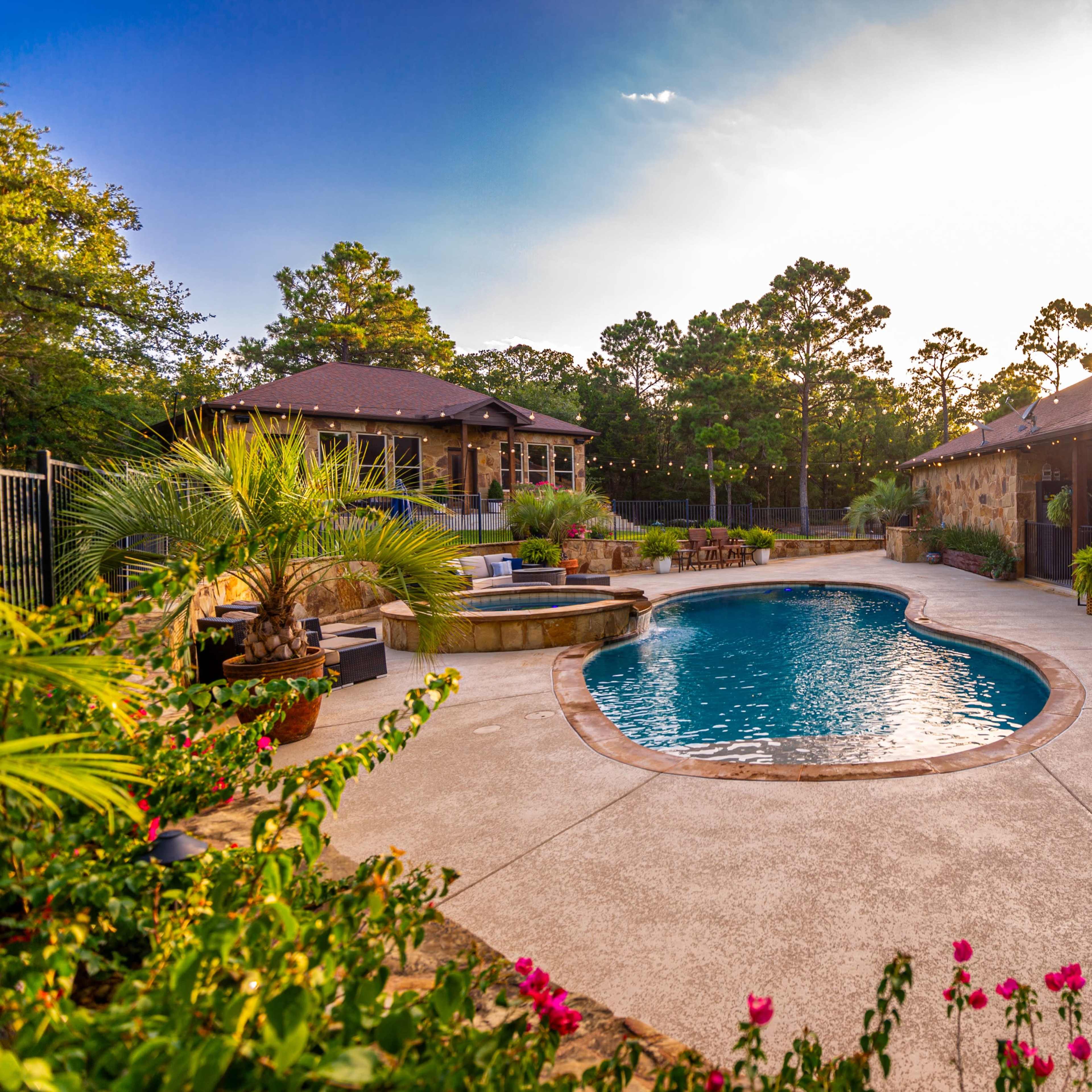 A landscaped backyard features a kidney-shaped swimming pool surrounded by seating areas and a hot tub, with lush greenery and flowers.
