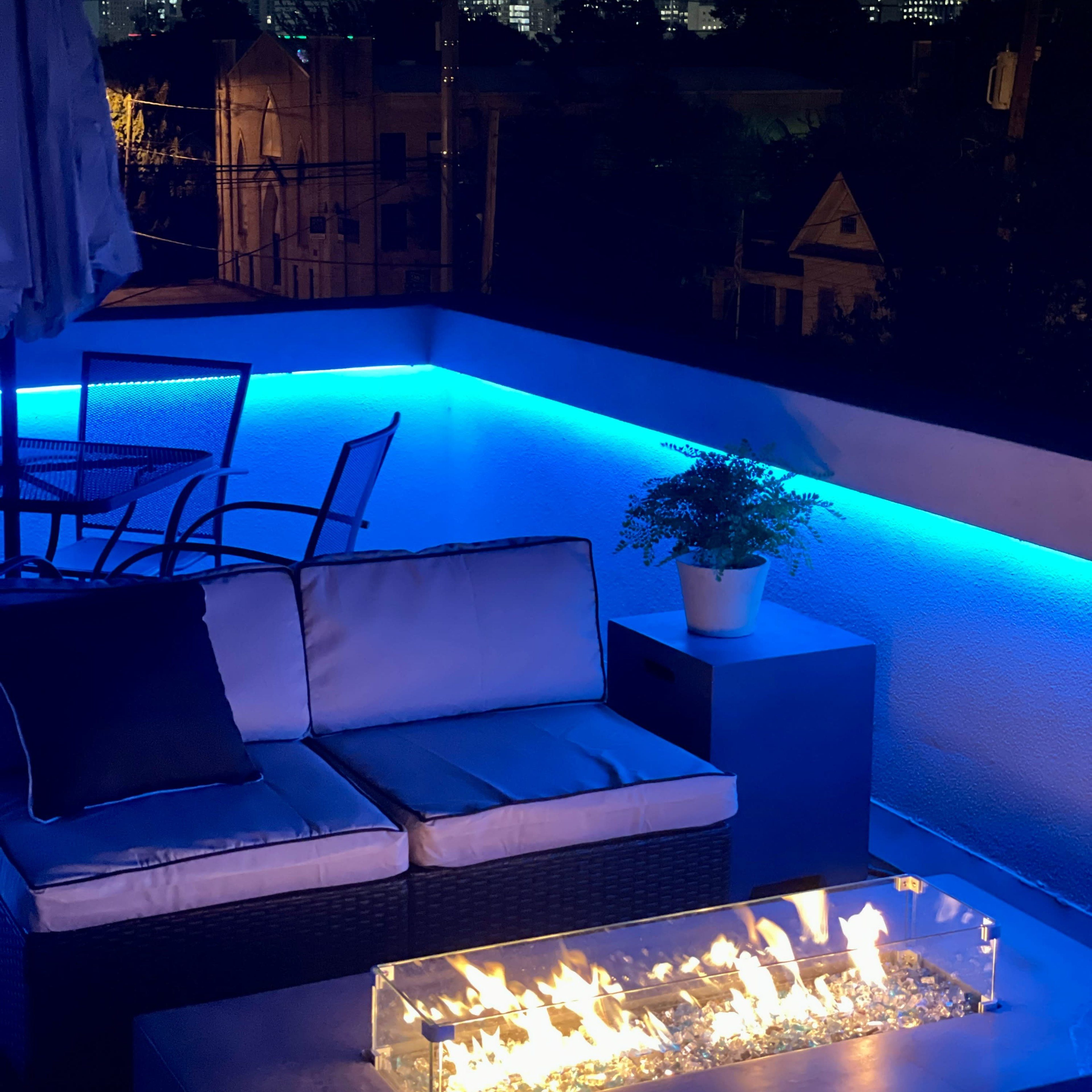 A rooftop patio features modern furniture with an outdoor fireplace and city skyline in the background at night.