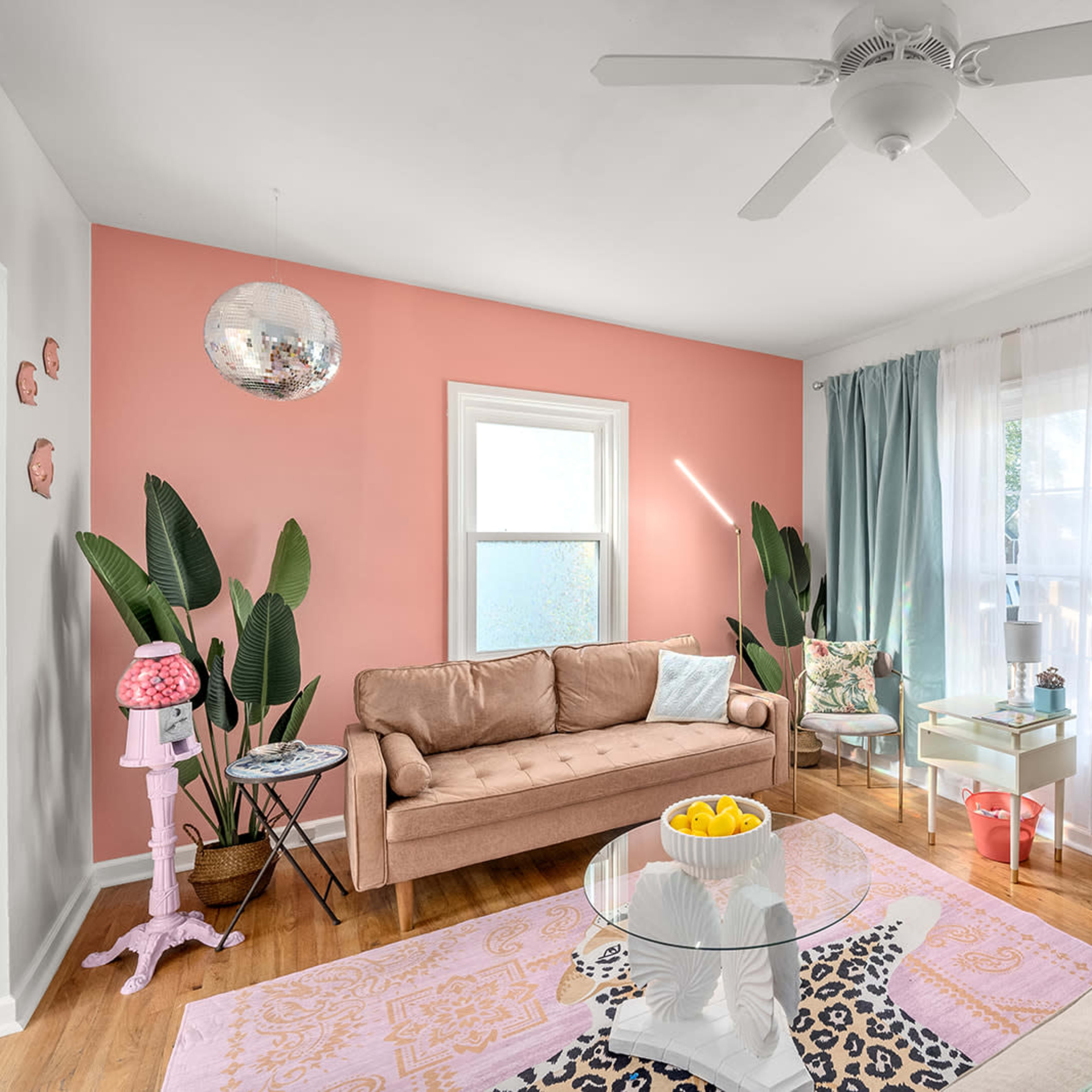 The living room features a pink accent wall, light-colored sofas, potted plants, and a large rug with a mix of patterns.