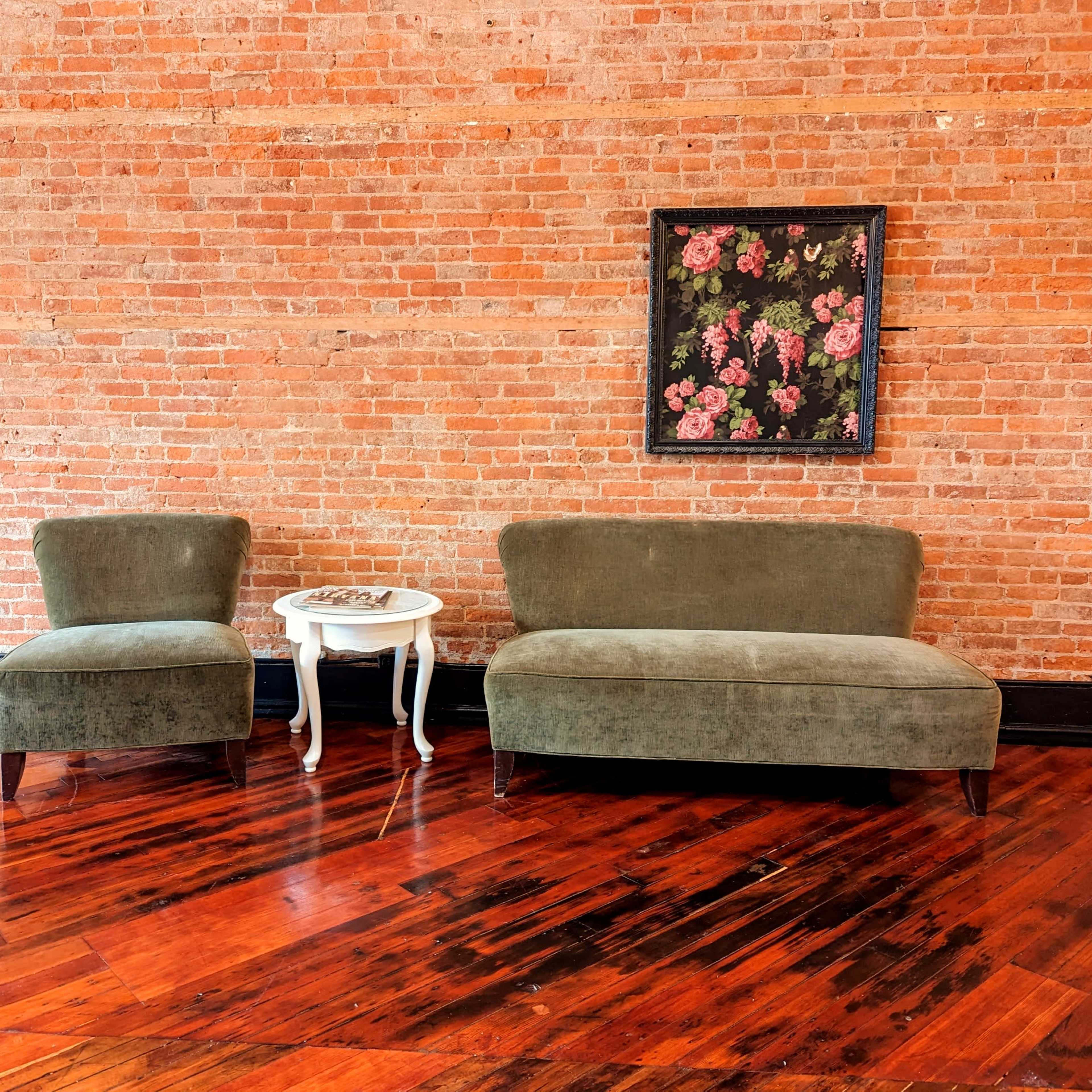 A seating area features two green upholstered chairs and a small white side table against a brick wall, with a floral painting above.