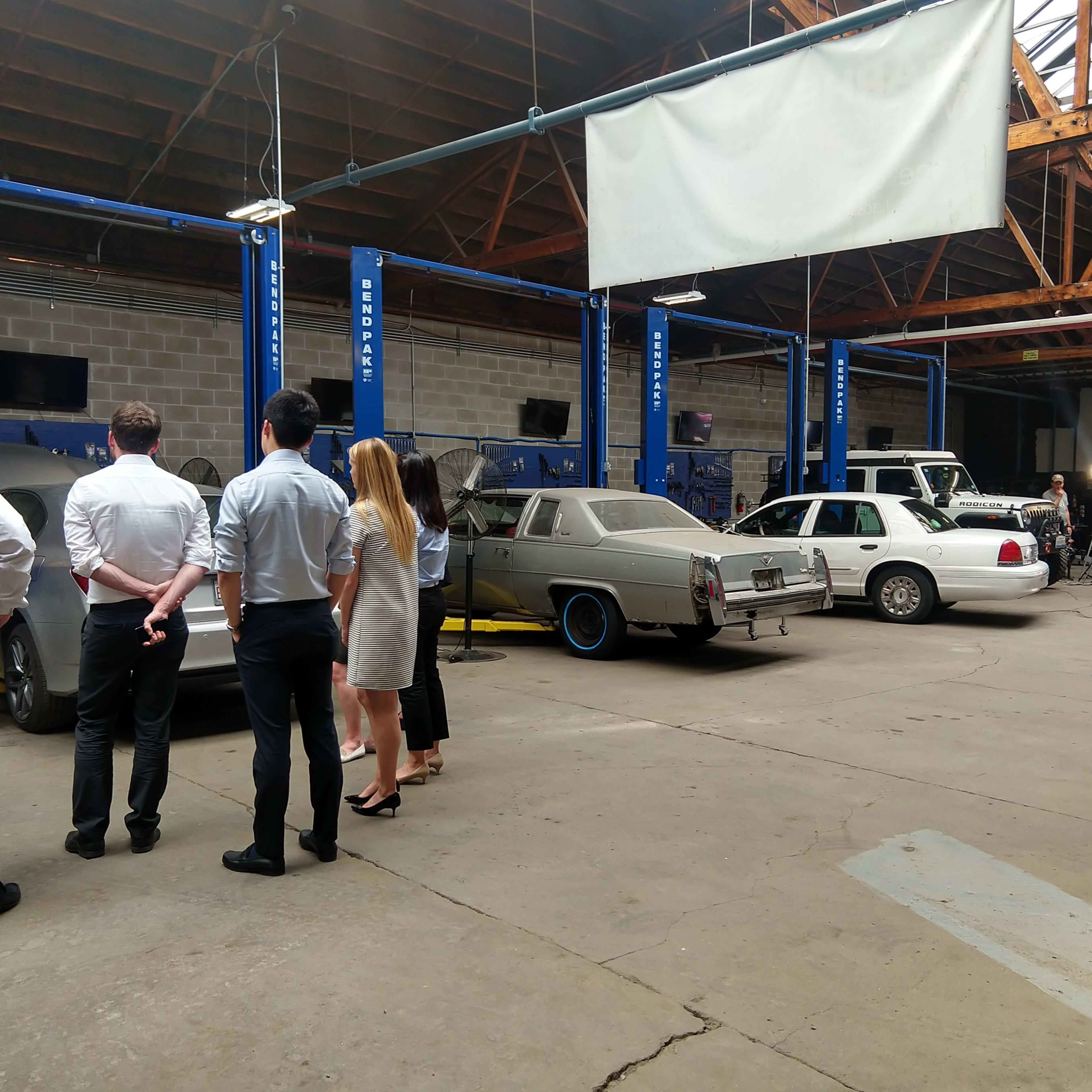 A group of people observing several cars inside a spacious automotive workshop with car lifts and exposed wooden beams overhead.