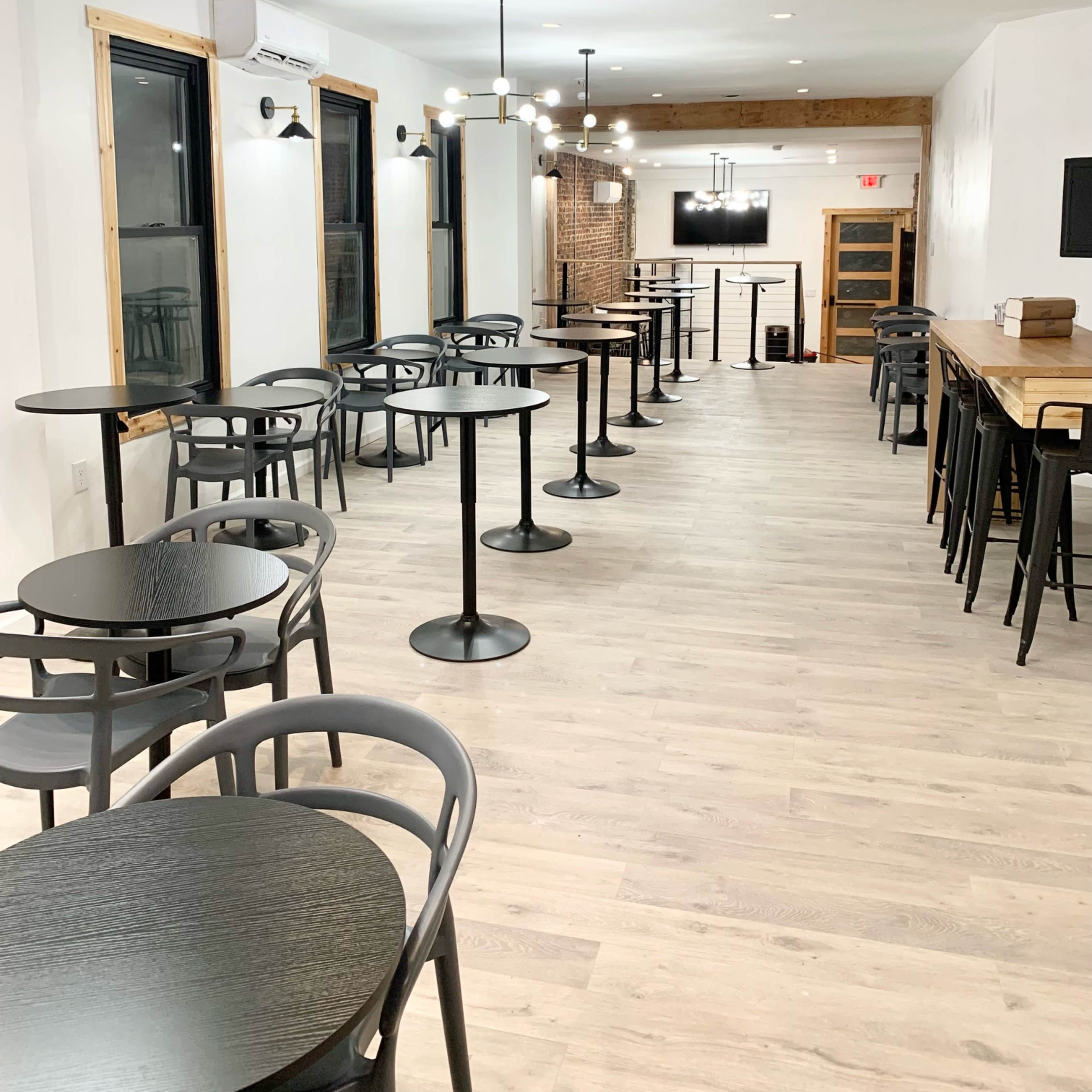 An empty café interior with several round tables and chairs arranged along the walls, and a long wooden counter at the end of the room.