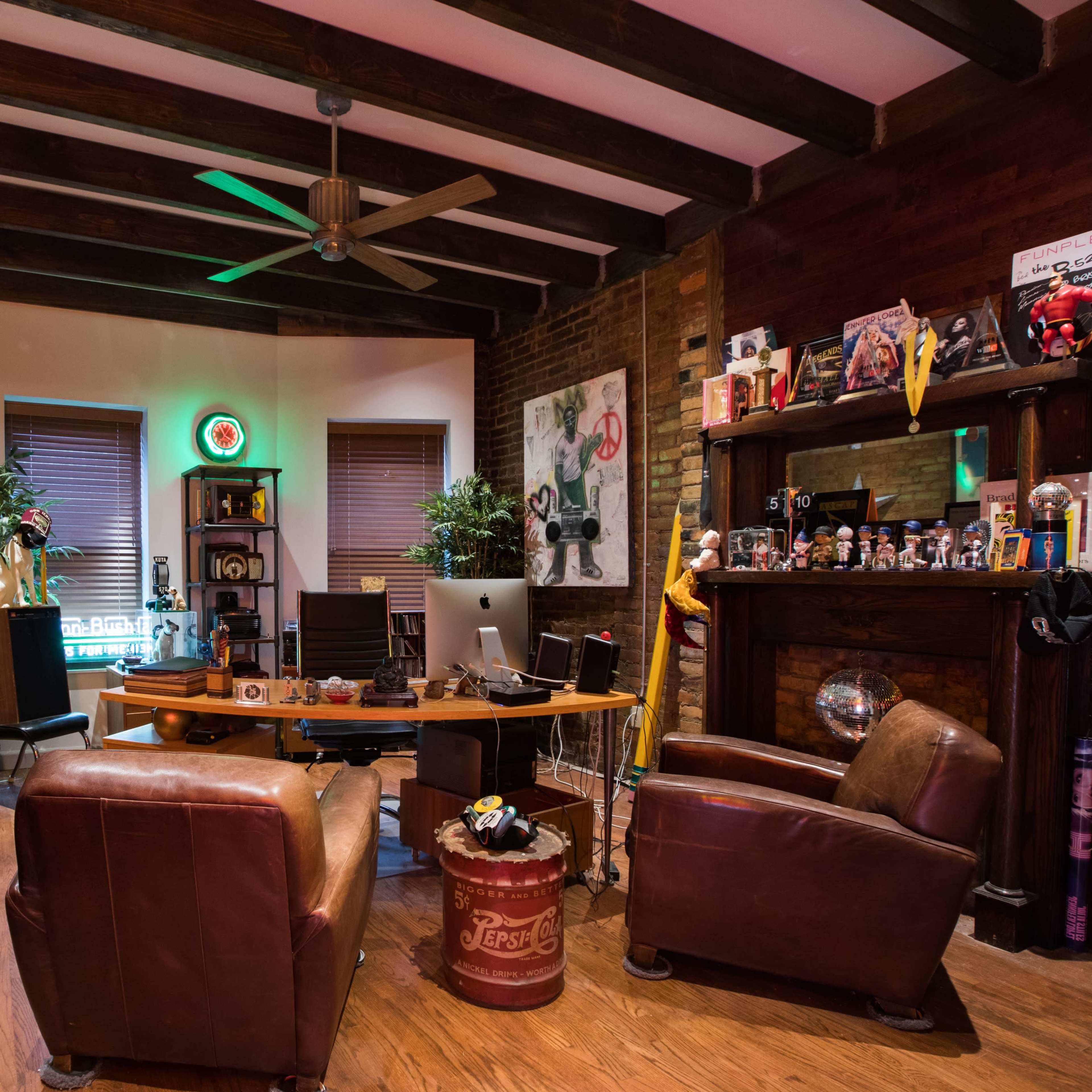 A cozy room features a wooden ceiling with beams, vinyl records on a side table, a desk with a computer, and various decorations including plants and collectibles.
