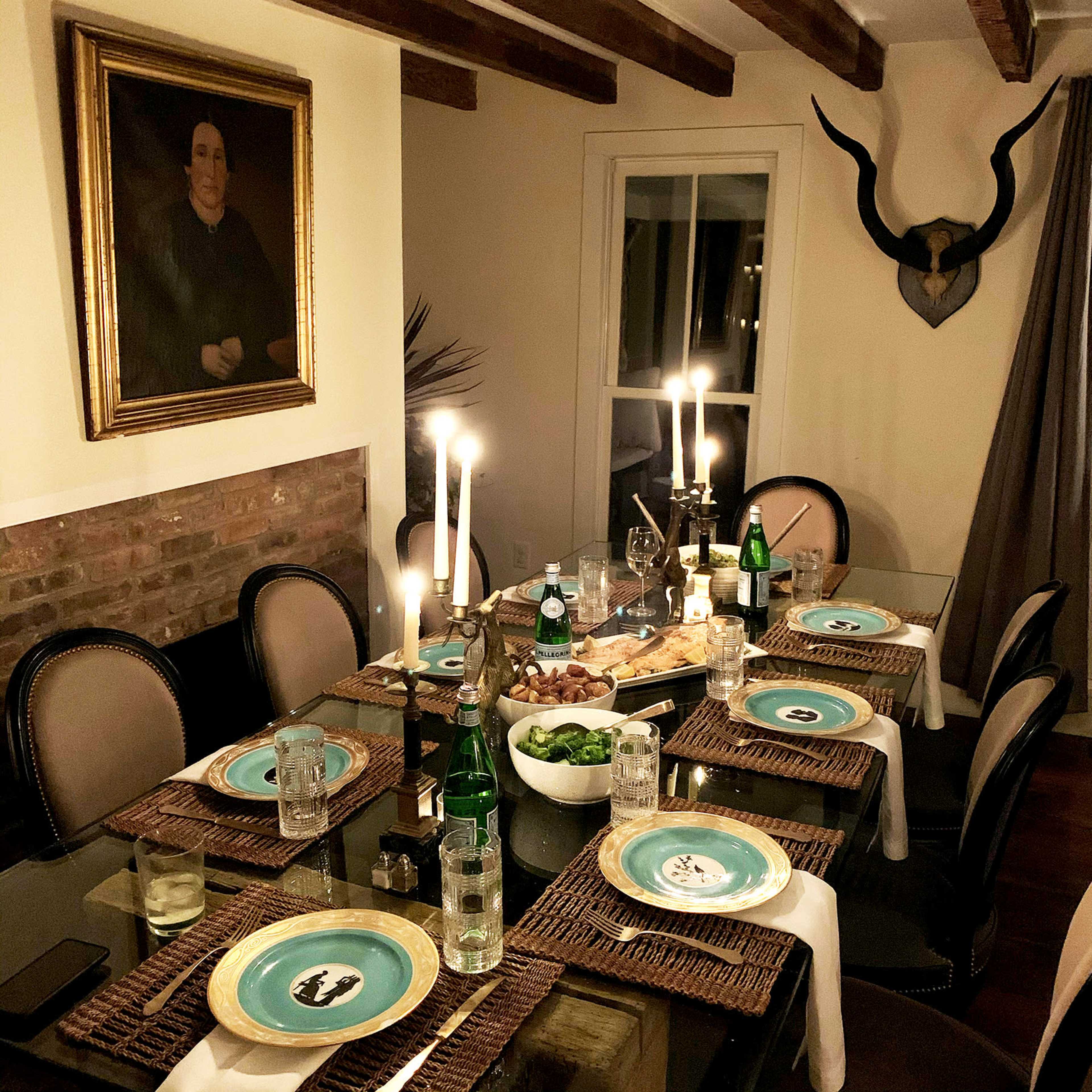 A dining table is set with blue plates, glasses of water, and candles, surrounded by a warm, inviting room featuring a large painting and decorative horns on the wall.