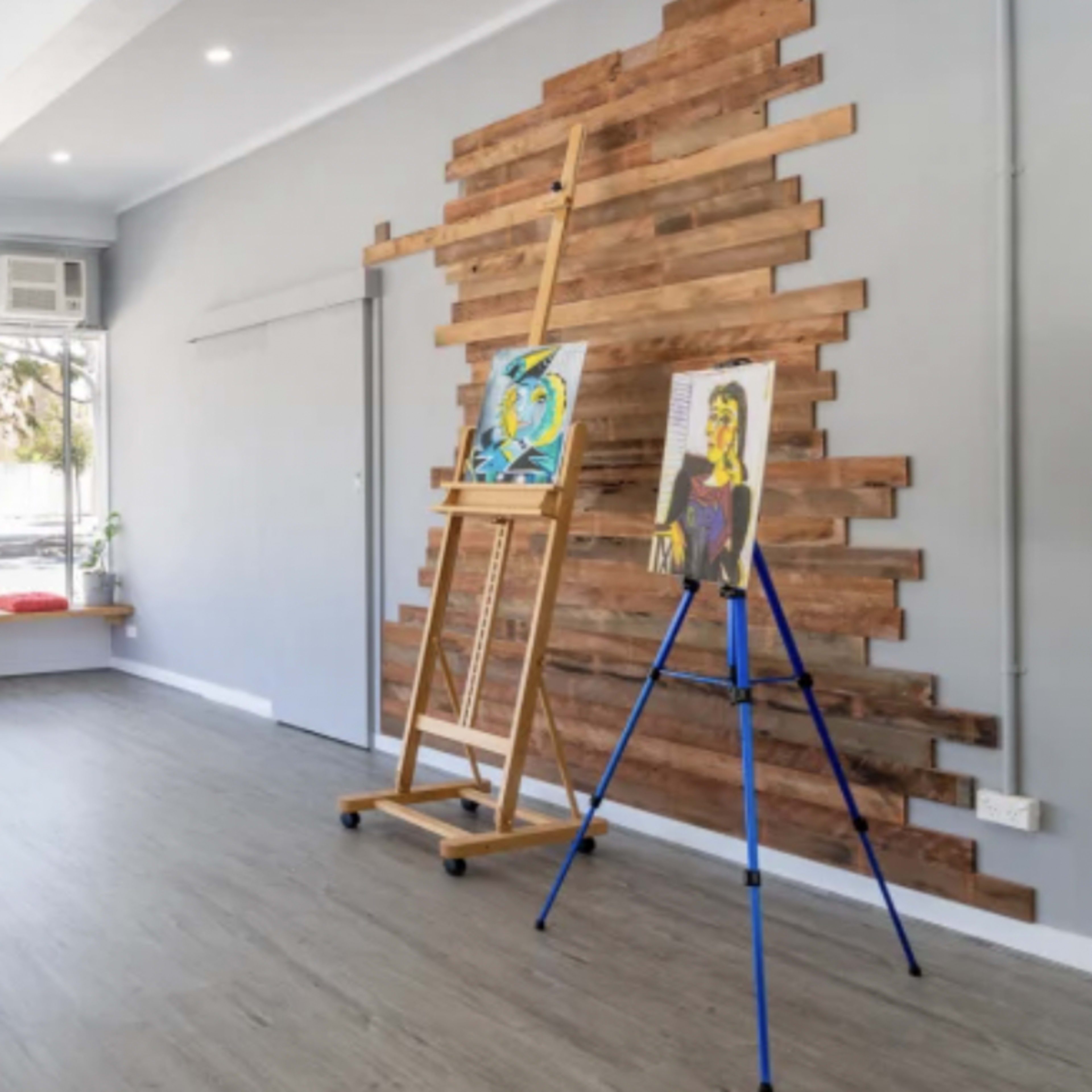 The interior of a spacious, well-lit art studio features wooden accents on the wall, an easel, and colorful cushions along the windowsill.