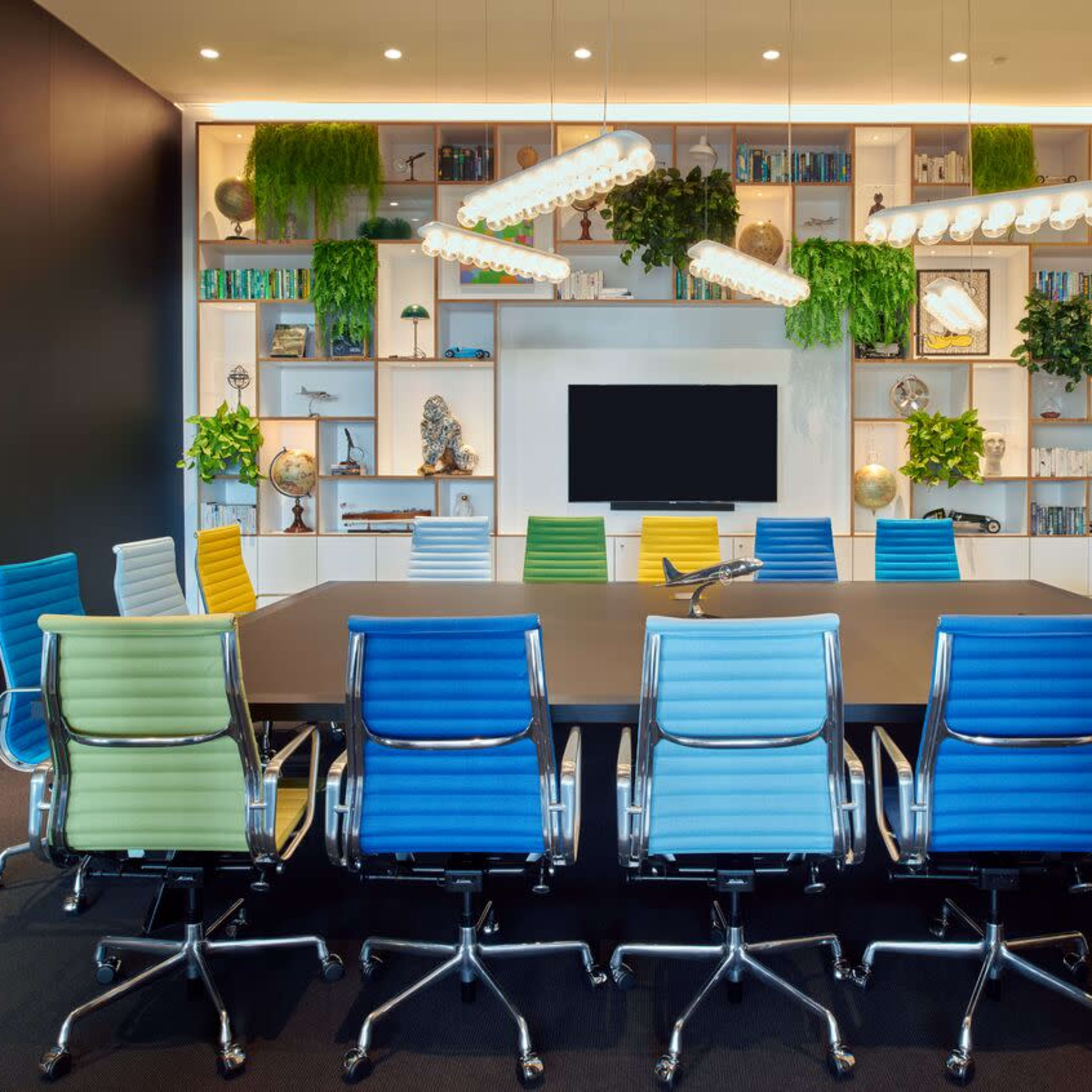 A modern conference room features a large table surrounded by colorful office chairs and a wall with shelves displaying plants and decorative items.