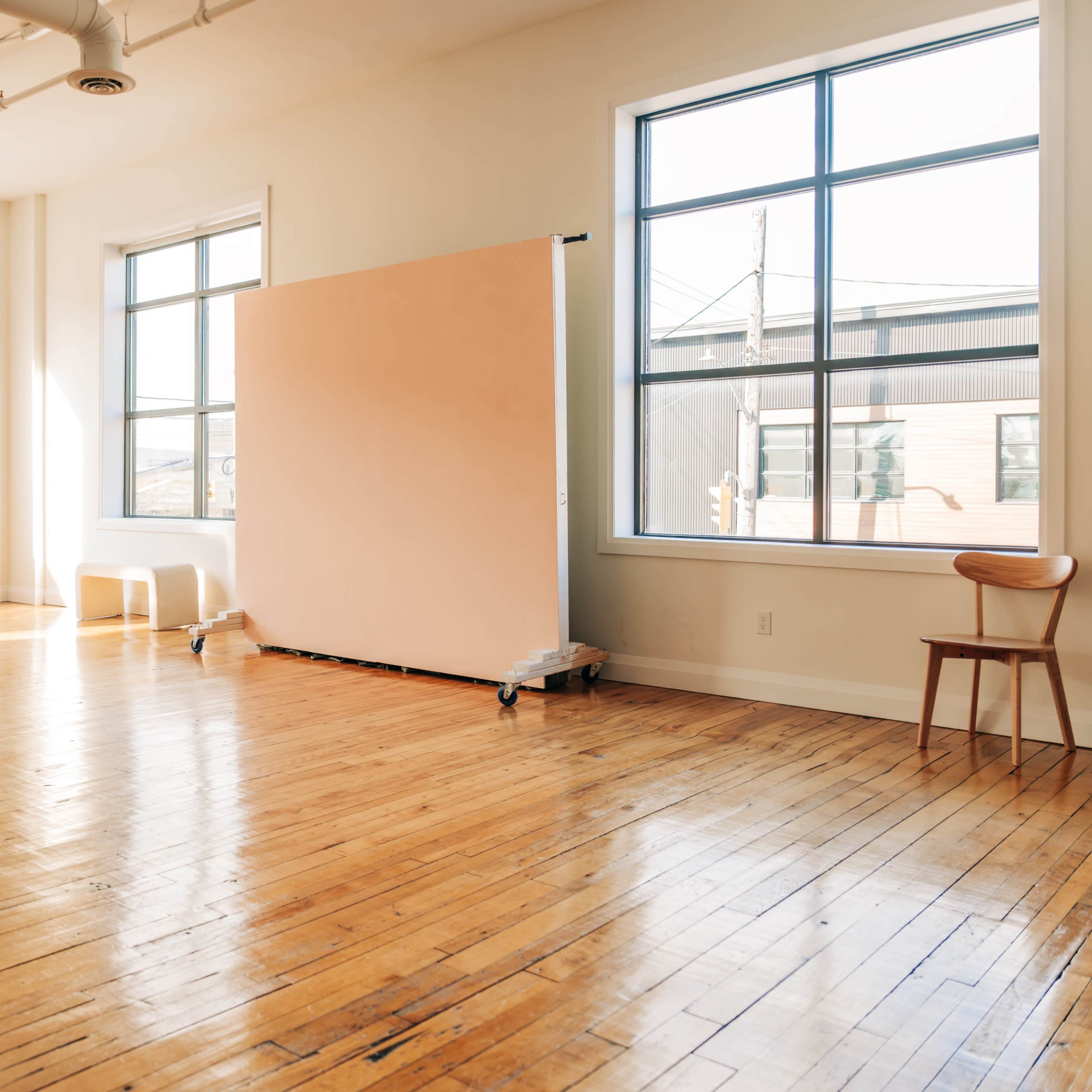 A bright, empty room features large windows, polished wooden floors, and a movable partition at one end.