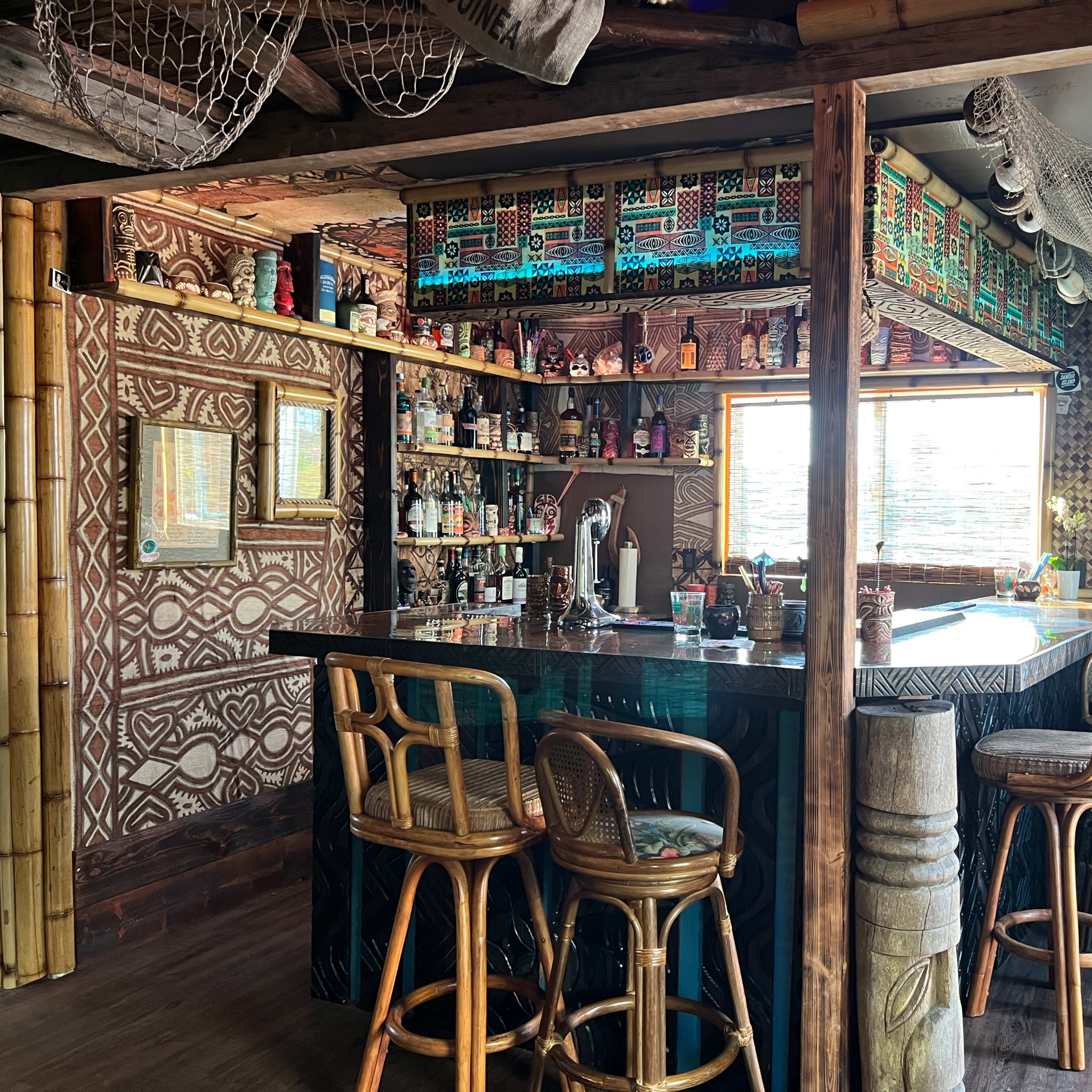 A tiki-themed bar with a bamboo frame, adorned shelves of colorful bottles, and a patterned wall design.