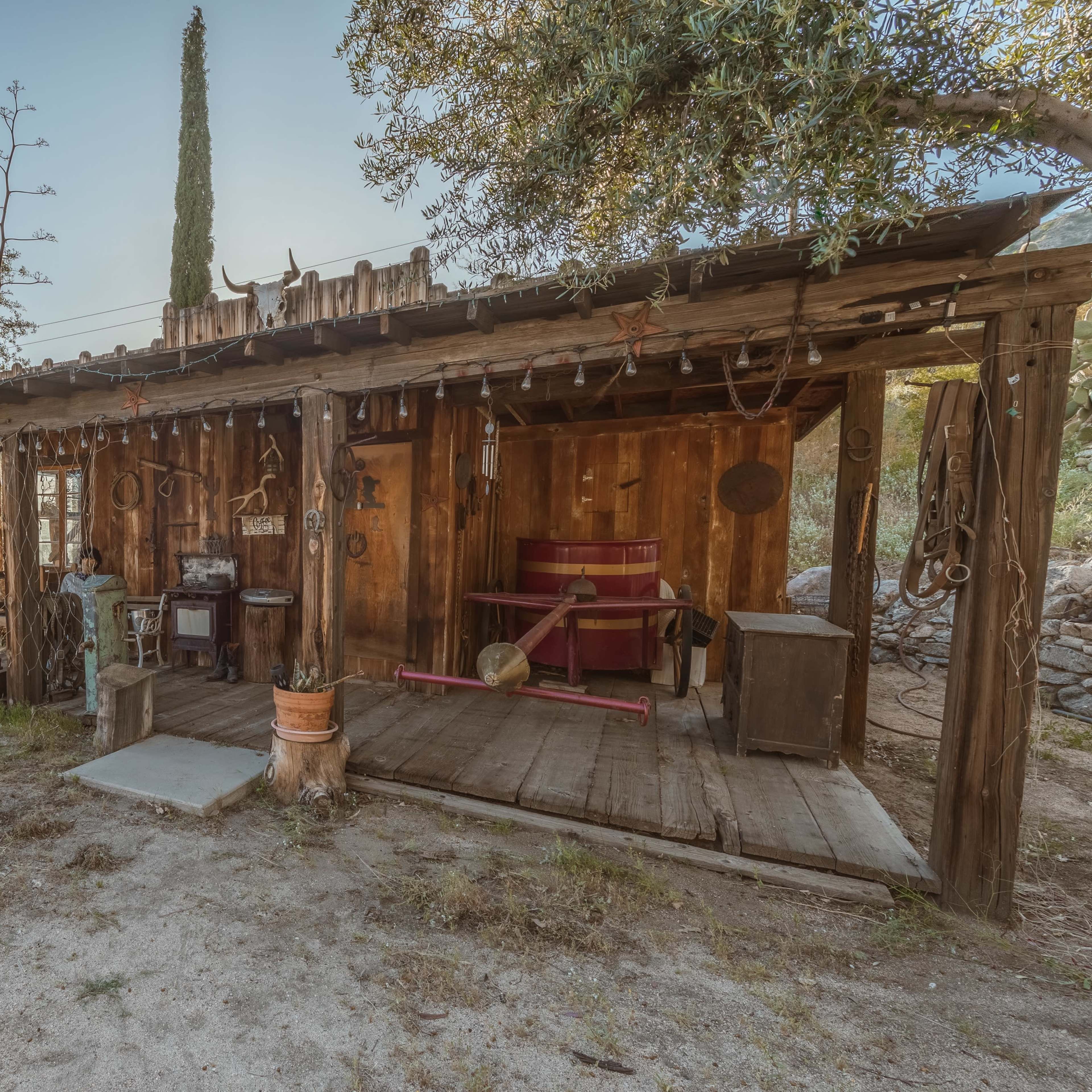A rustic wooden structure surrounded by natural vegetation, featuring a porch with various tools and a wagon wheel in the foreground.