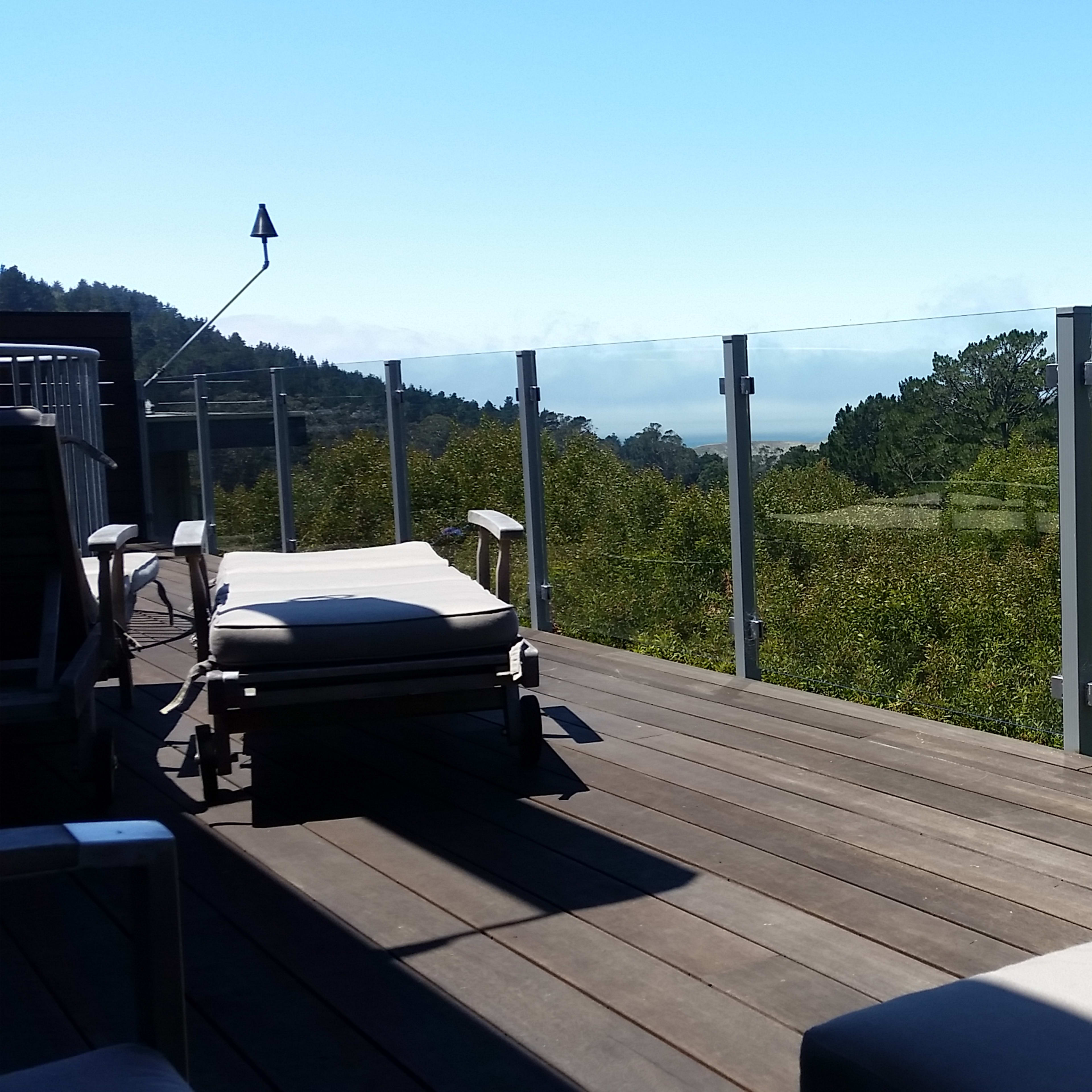 A wooden deck with large glass panels overlooks a green landscape and distant hills under a clear blue sky.