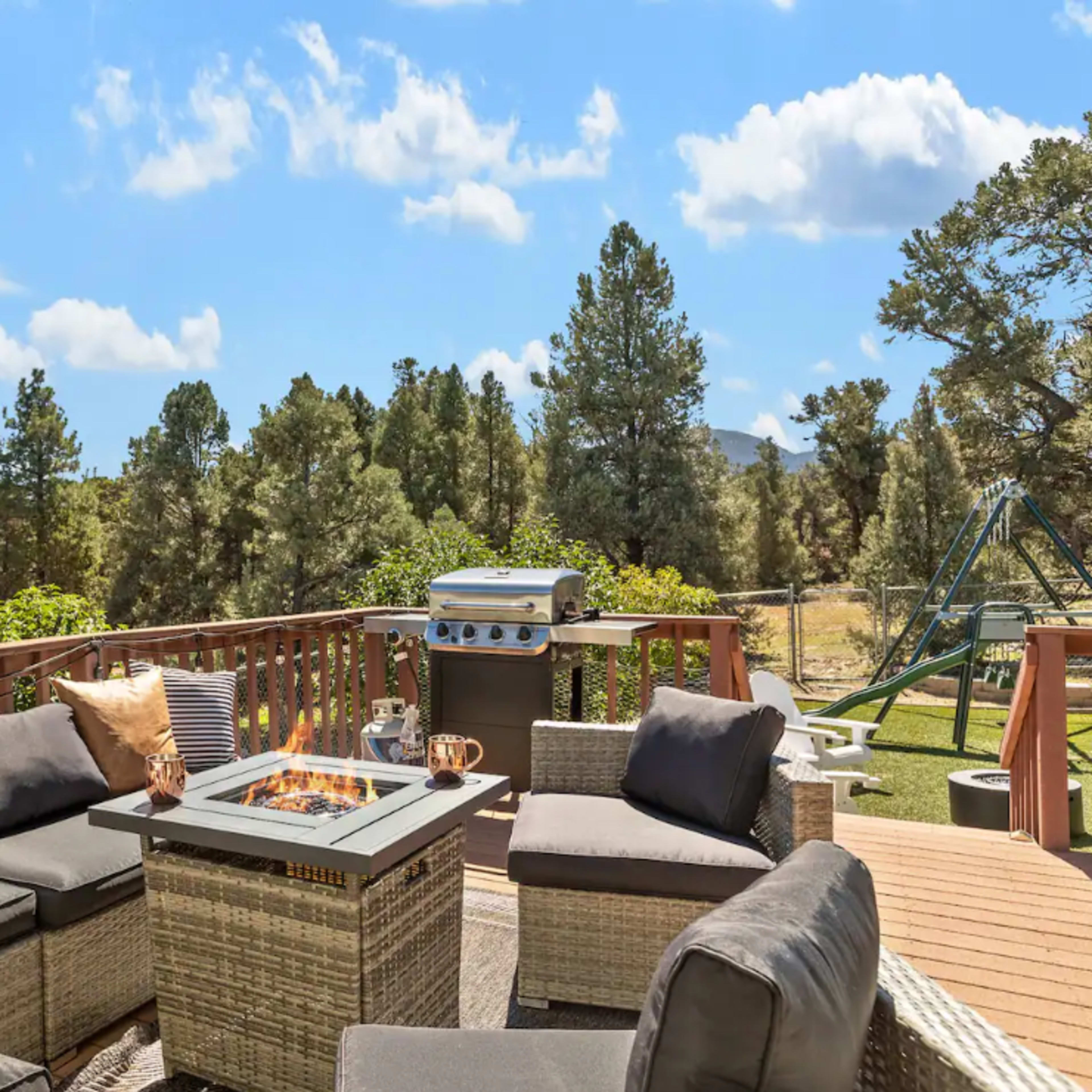 A patio area with comfortable seating, a fire pit table, a grill, and a view of lush trees and blue sky in the background.