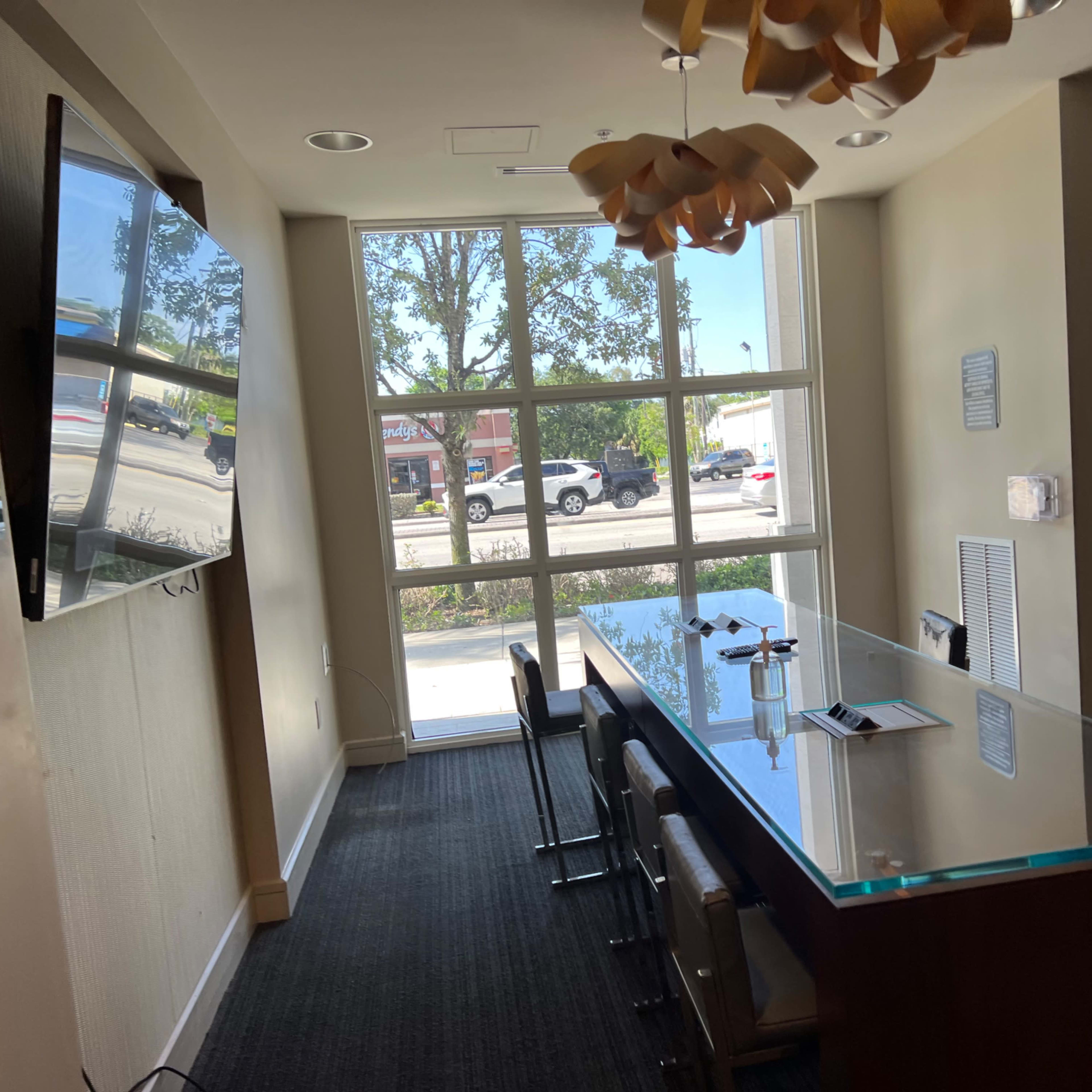 A meeting room features a long glass table, chairs, a television on the wall, and large windows providing a view of the street outside.