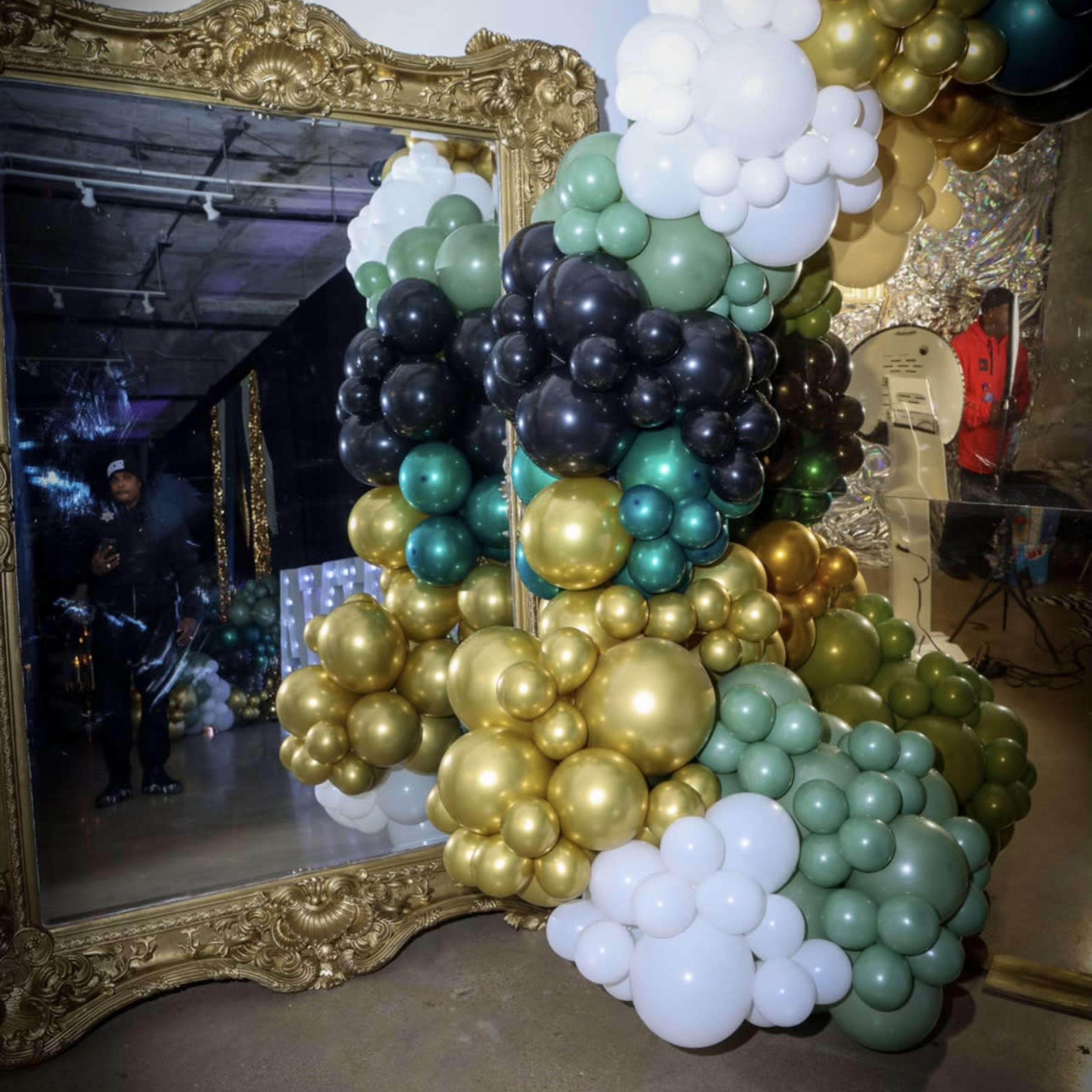 A large arrangement of colorful balloons in black, gold, green, and white surrounds an ornate mirror in a decorated space.
