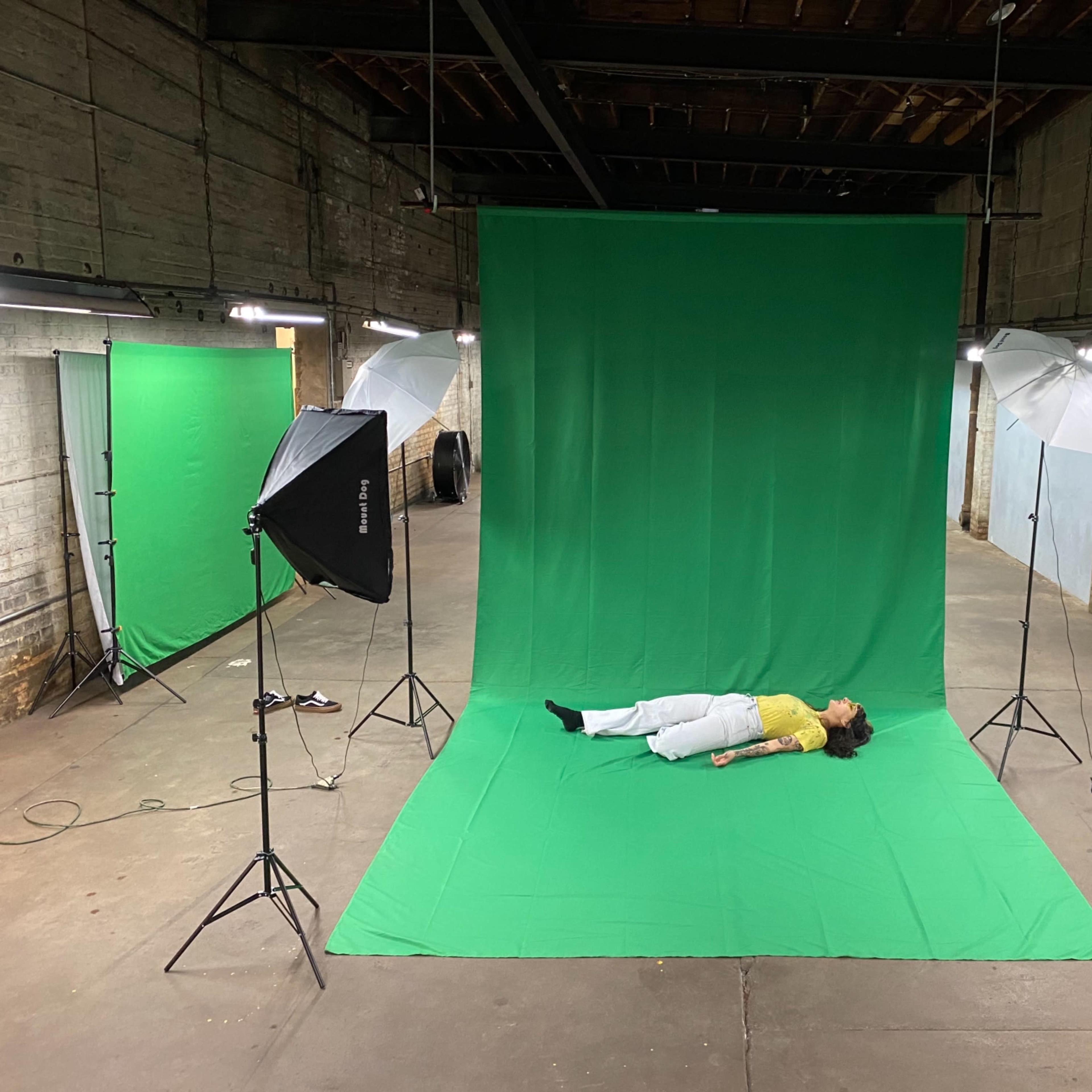 A person lies on a green backdrop in a dimly lit studio, surrounded by umbrella lights and additional green screens.