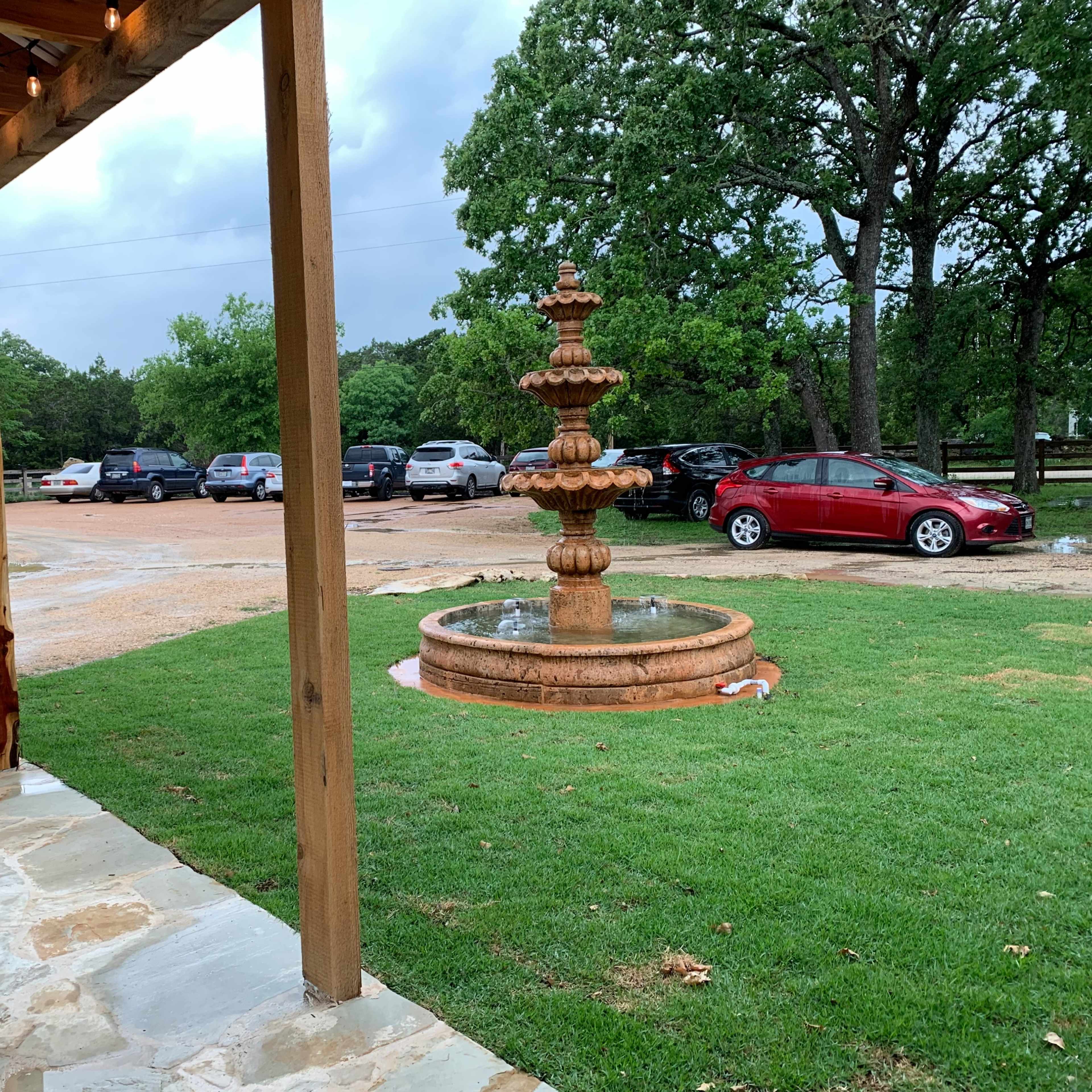 A fountain stands in the foreground as two people watch a parking area filled with cars under overcast skies.