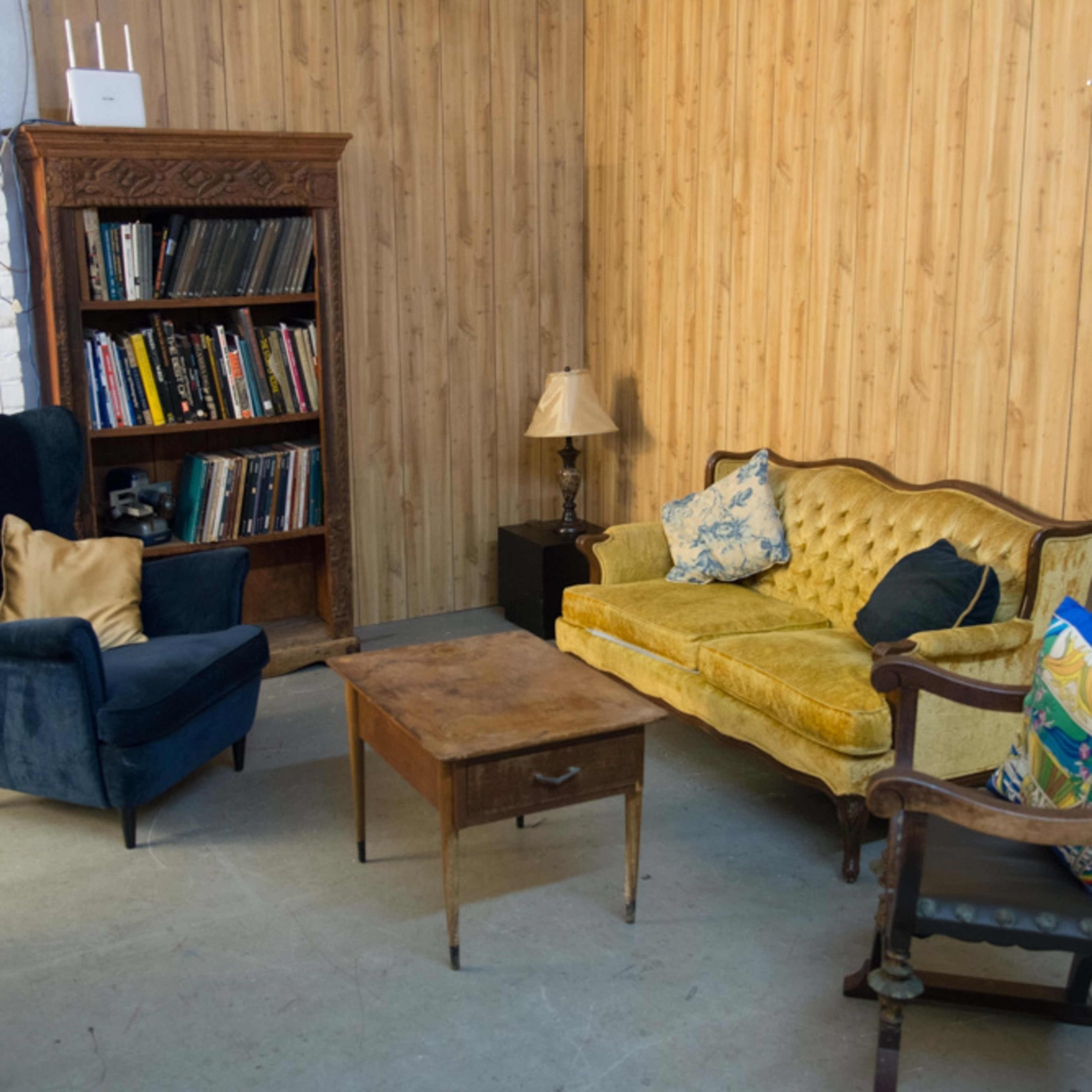 A small living area with two upholstered chairs, a yellow sofa, a wooden coffee table, and a bookcase against a wooden-paneled wall.