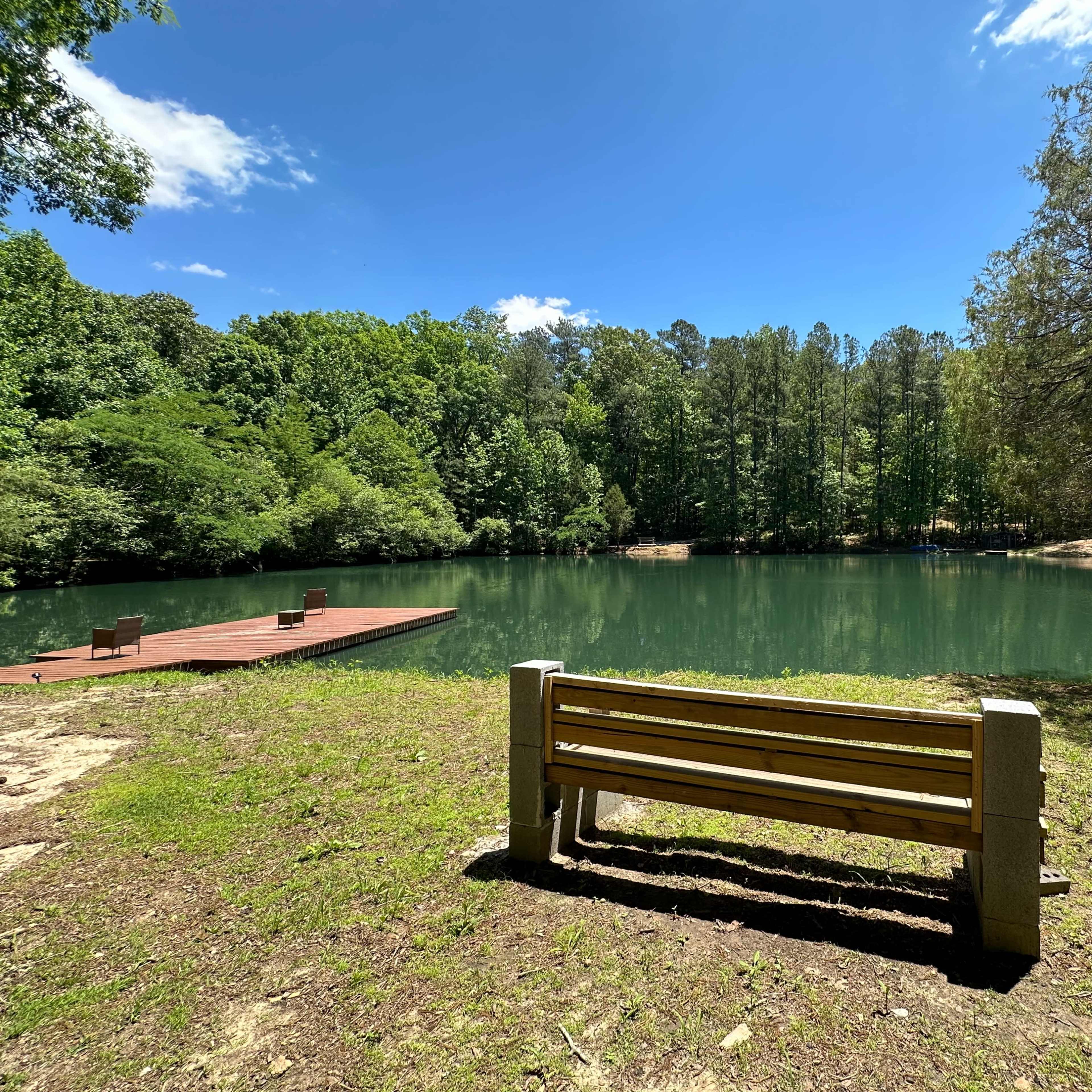 A wooden bench faces a calm, green lake surrounded by forested land and a small dock in the background.