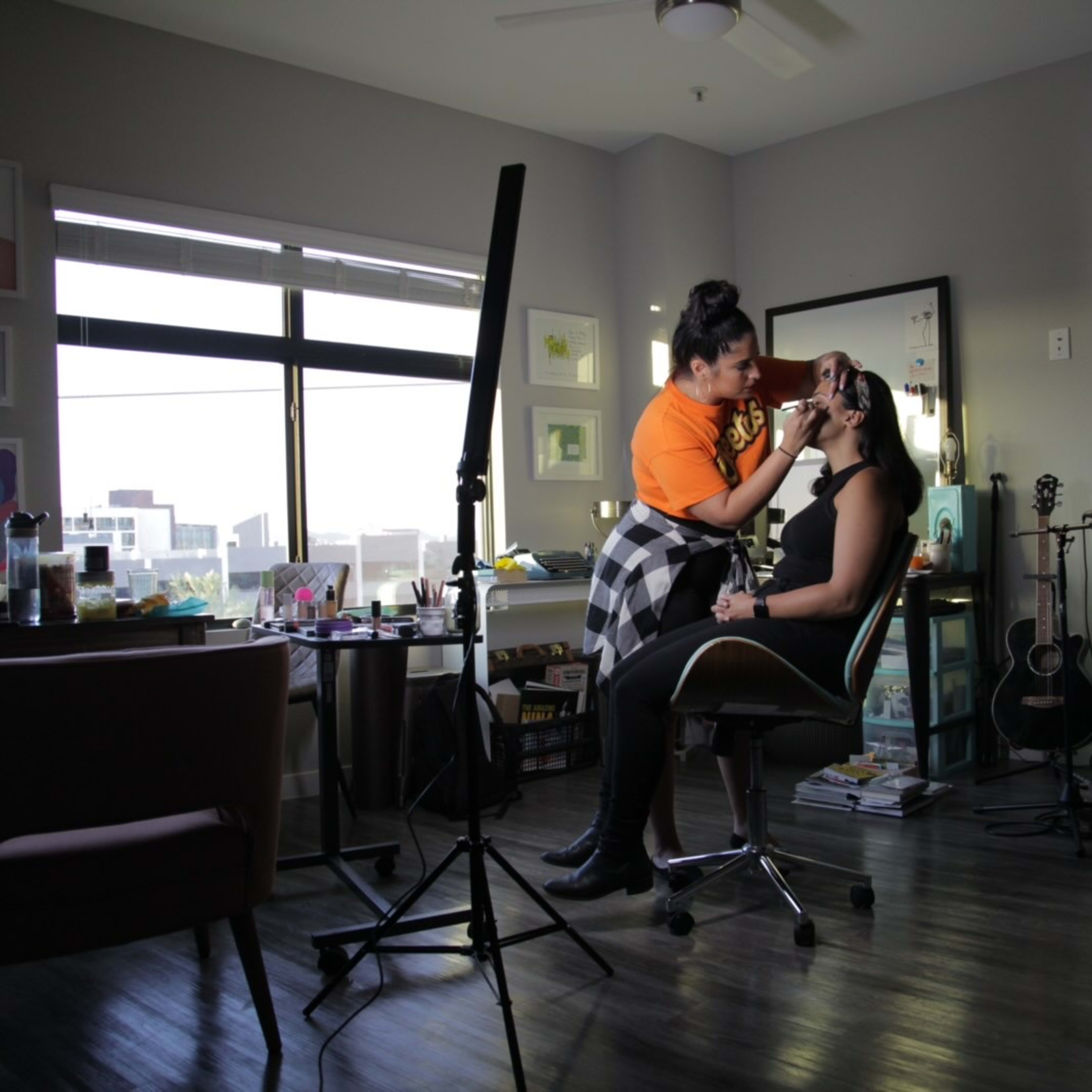 A makeup artist applies makeup to a client in a well-lit room with studio equipment and a desk filled with beauty products.