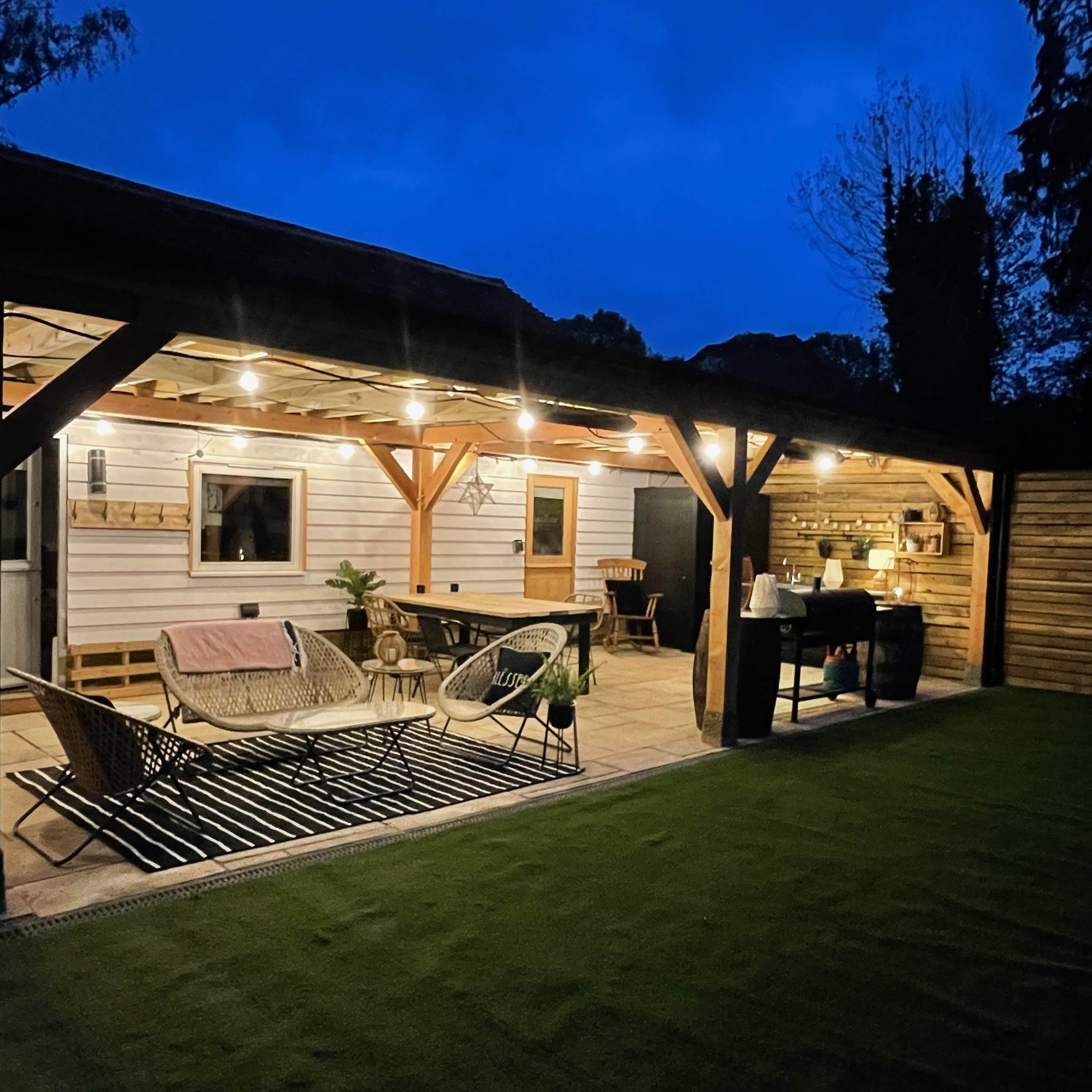 A well-lit outdoor patio featuring a seating area with two lounge chairs and a table, surrounded by lush greenery under a twilight sky.