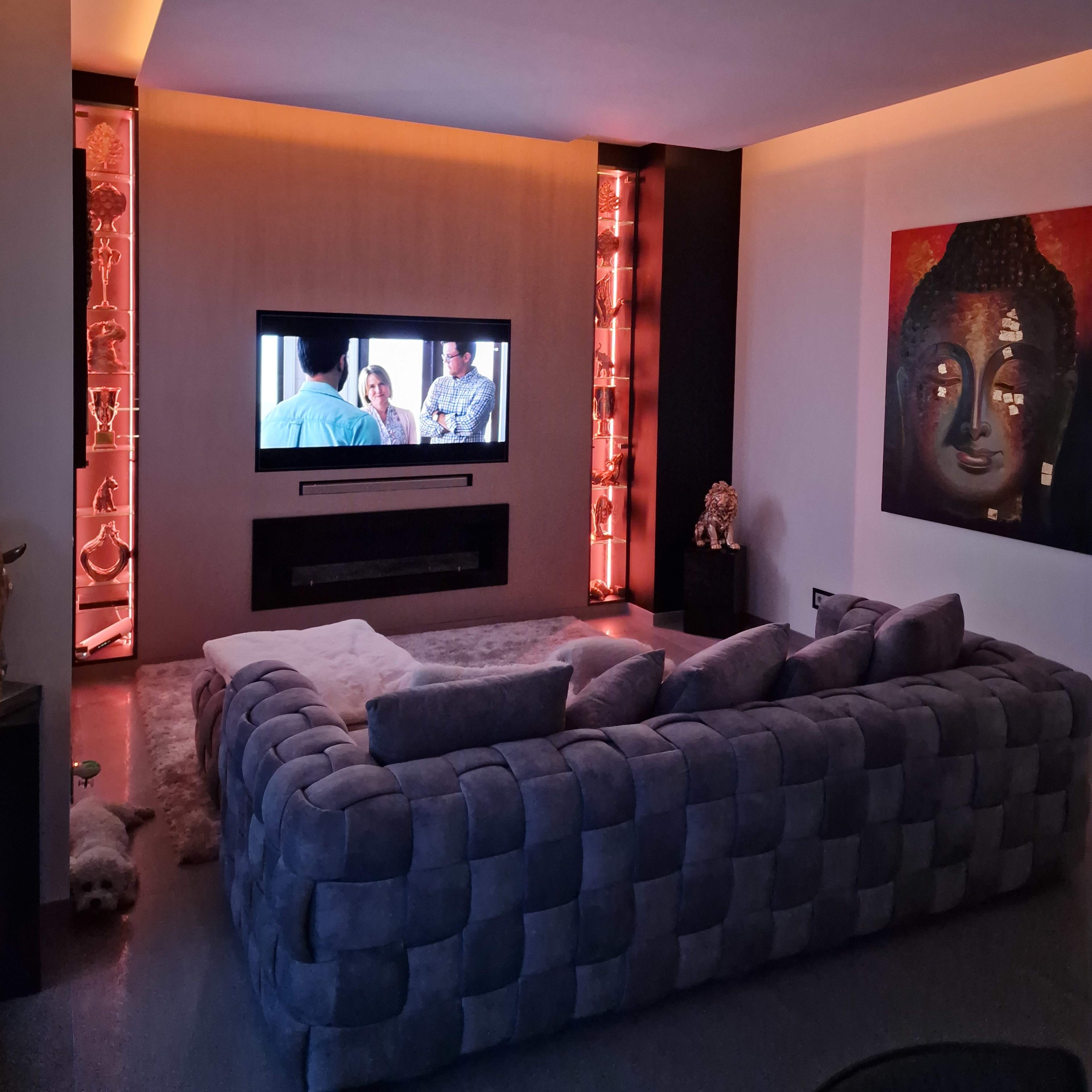 A modern living room with a large gray sofa facing a wall-mounted TV, surrounded by decorative shelves with ambient lighting and a large Buddha painting on the adjacent wall.