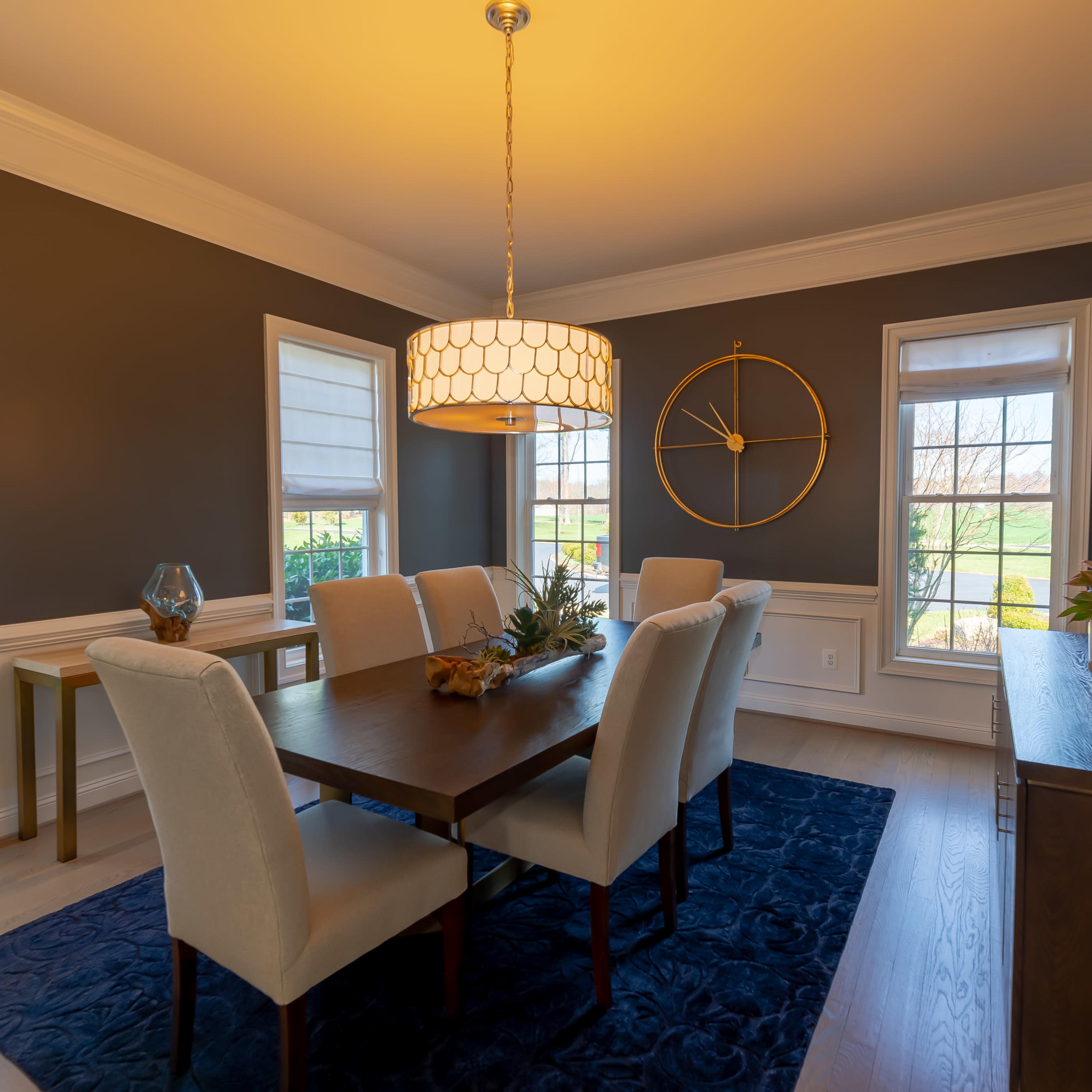 A dining room features a wooden table surrounded by six upholstered chairs, with a pendant light overhead and windows allowing natural light to enter.