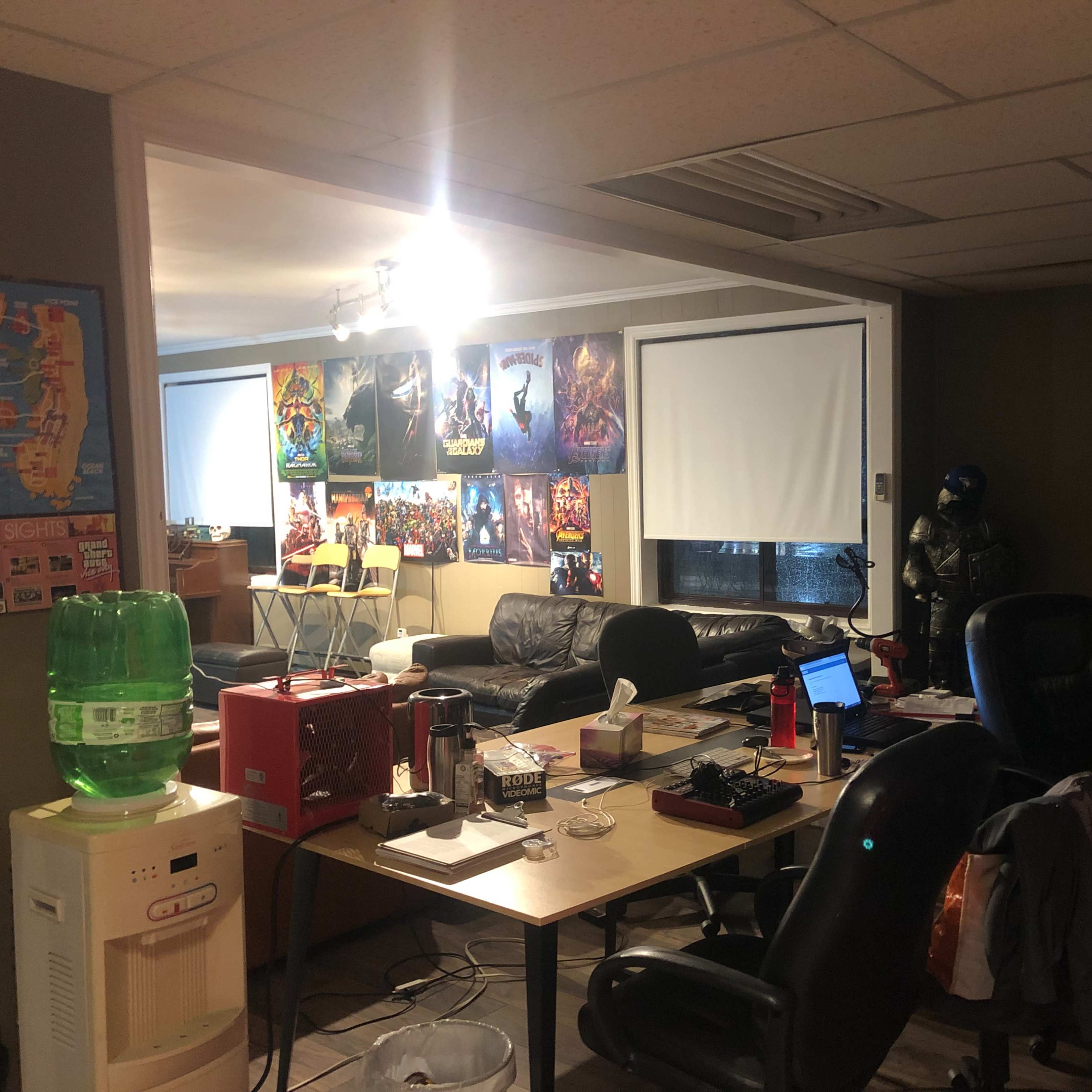 A cluttered office space features a desk with multiple chairs, a couch, posters on the walls, and various equipment such as a water cooler and speakers.