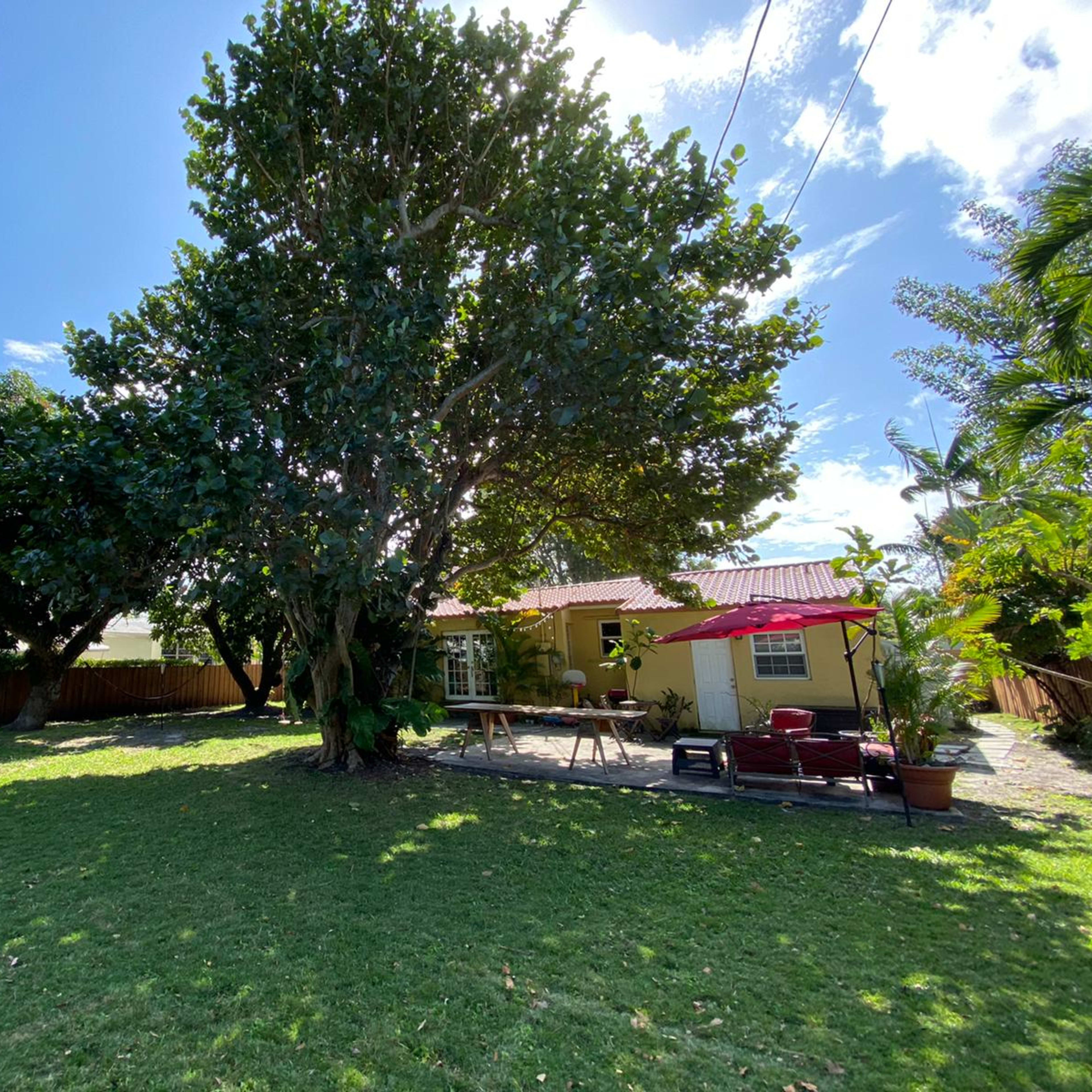 A house with a yellow exterior is located in a grassy yard under a large tree, with outdoor furniture arranged nearby.