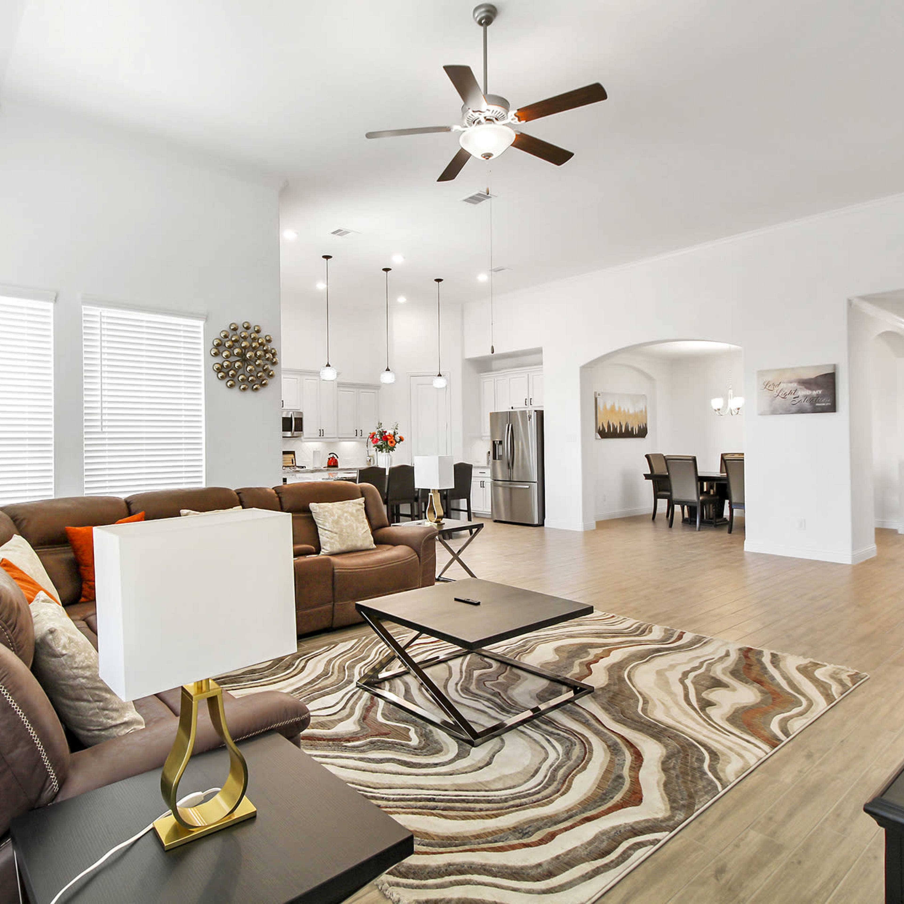A spacious living room with a brown sectional sofa, a coffee table, and a large window allowing natural light, leading to an open kitchen and dining area.