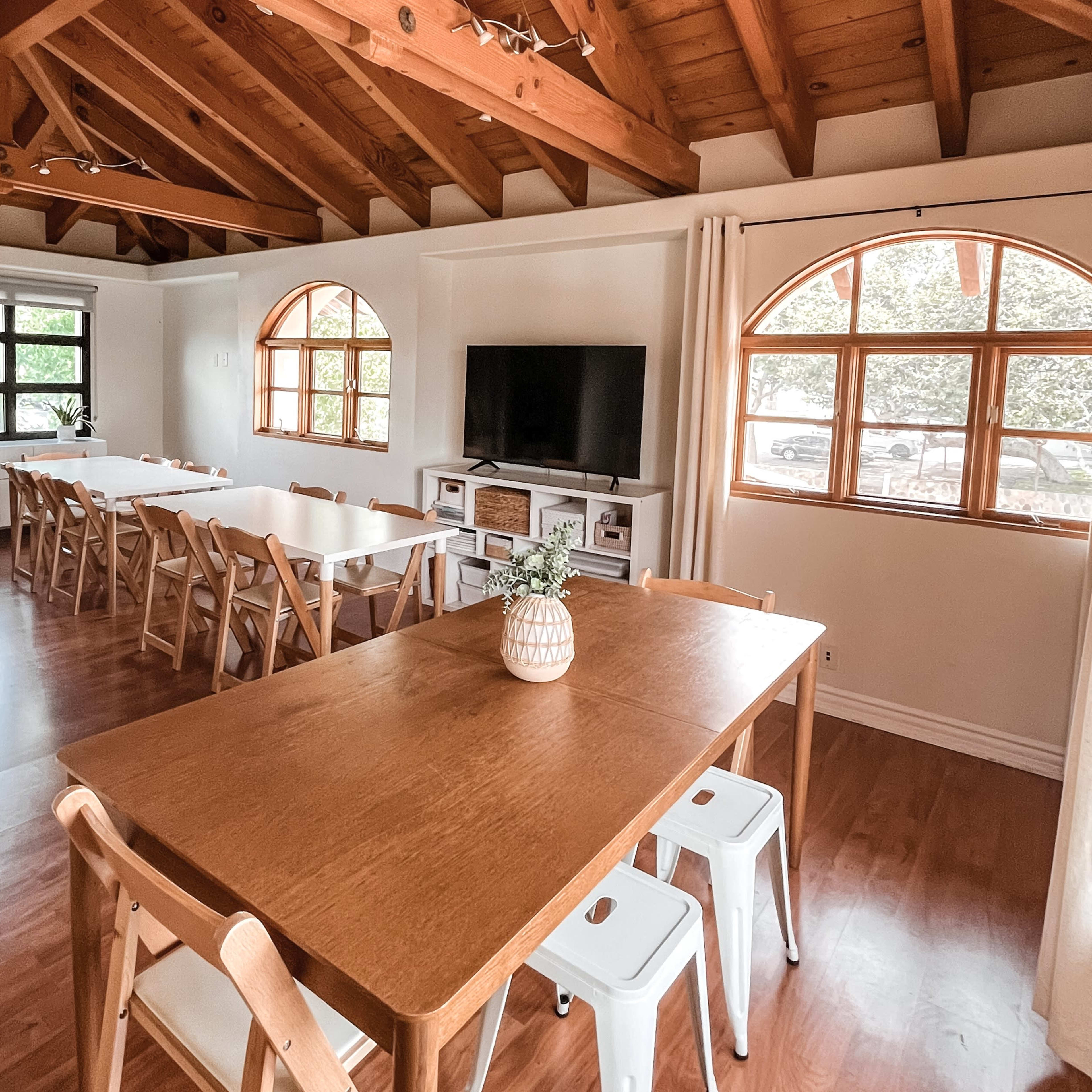 A spacious interior with wooden beams, two large dining tables, and a television against a wall, illuminated by natural light through arched windows.