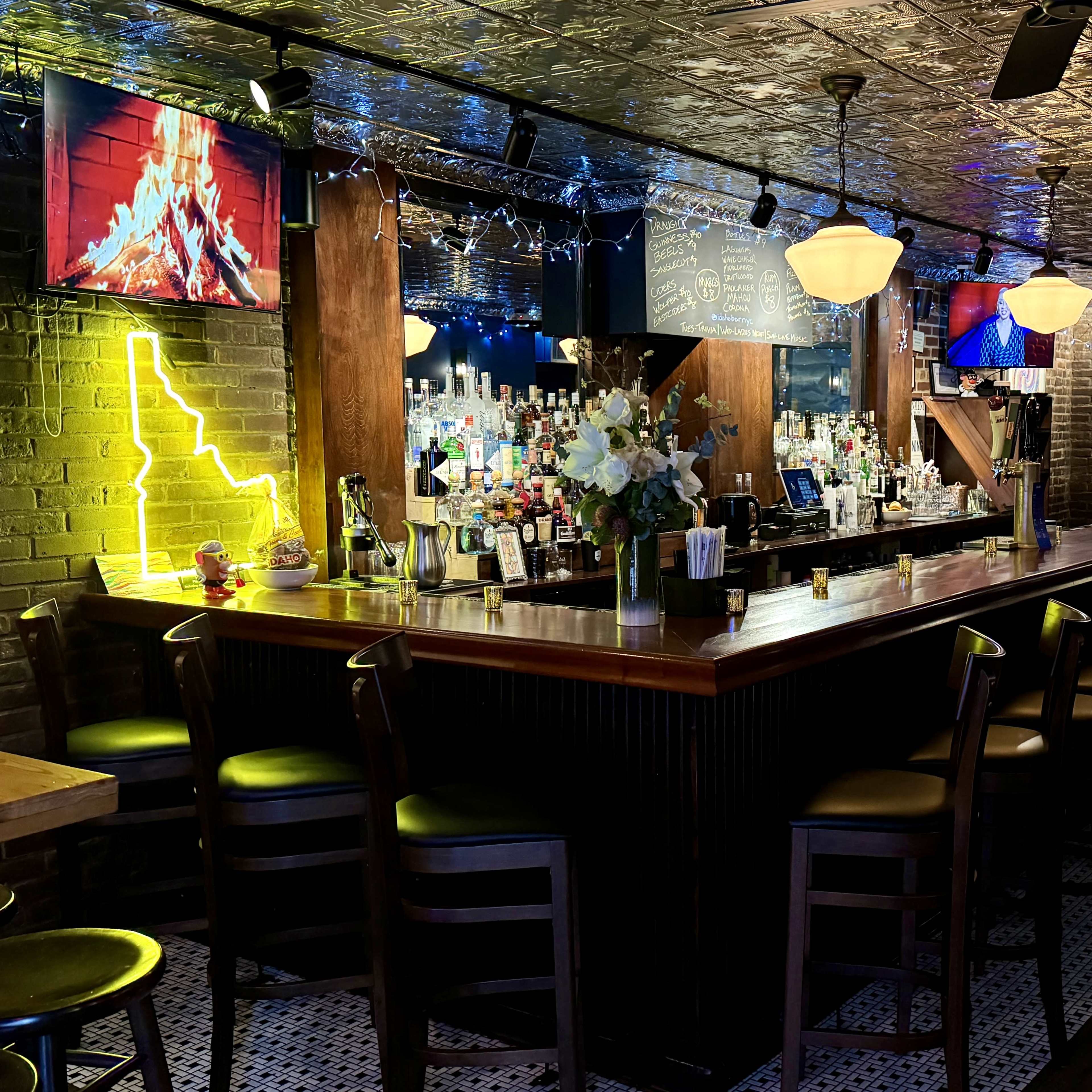 A dimly lit bar area with a long wooden counter, shelves stocked with drinks, and multiple flat-screen TVs mounted on the walls.