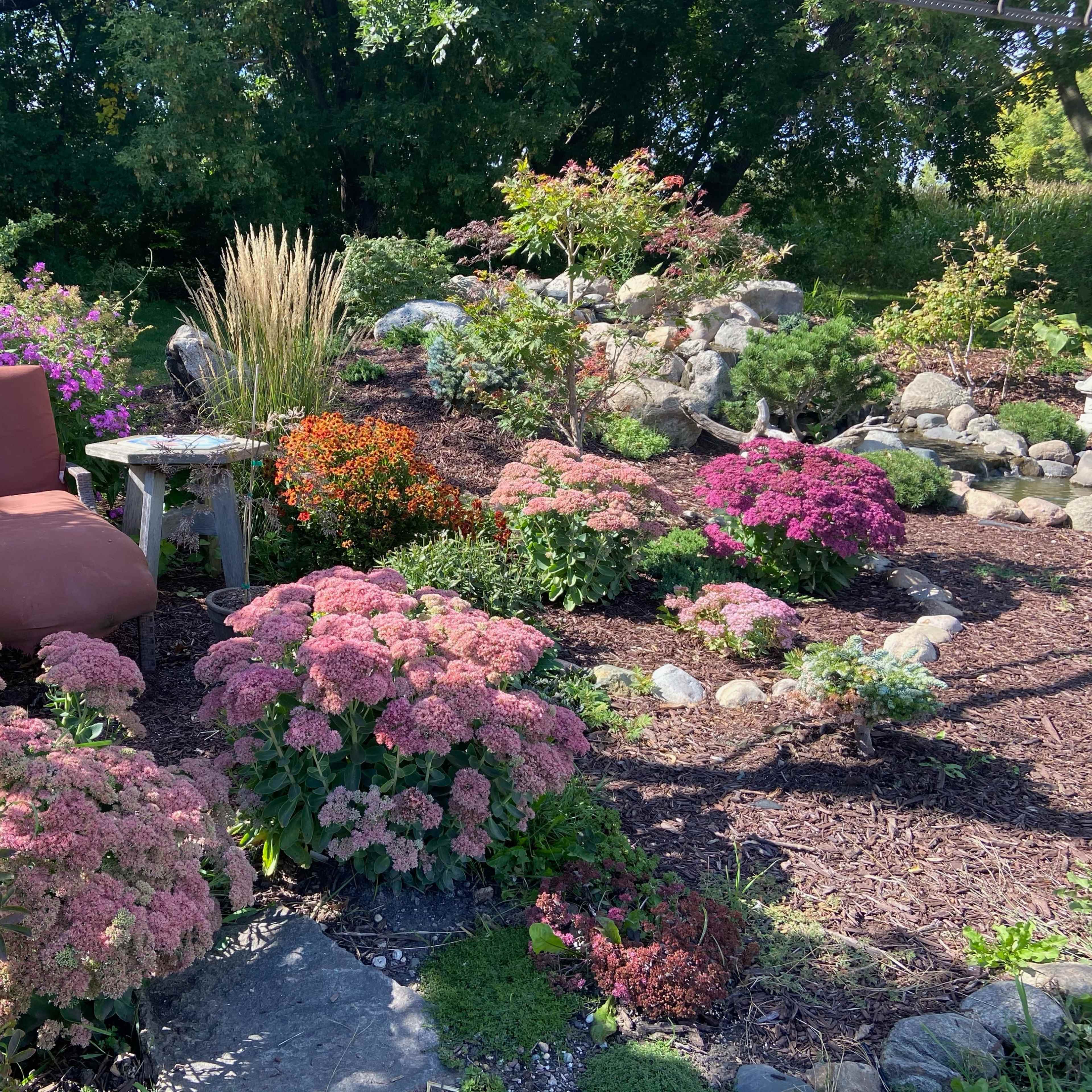 A cozy garden scene features a large chair amidst vibrant flowers and decorative rocks, with a small table nearby.